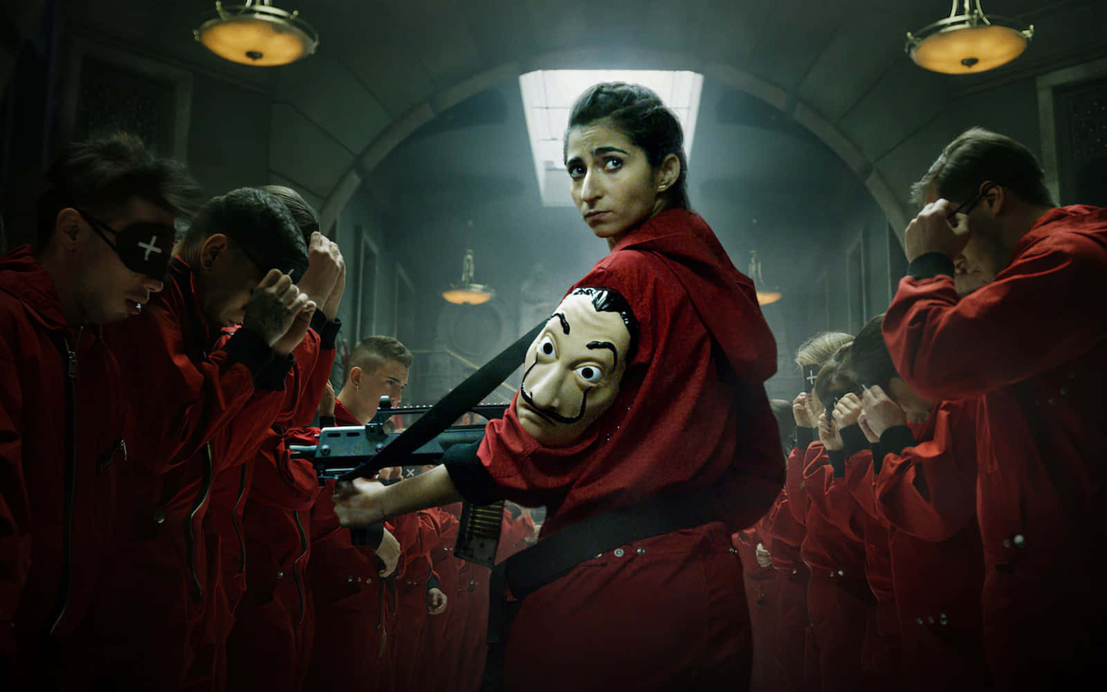 Money Heist Mask 桌布 壁紙 — 來自《魷魚遊戲》系列的緊張場景，一名戴面具的玩家身穿紅色連身衣手持步槍，其他穿紅色制服的人在昏暗的房間裡遮住眼睛。