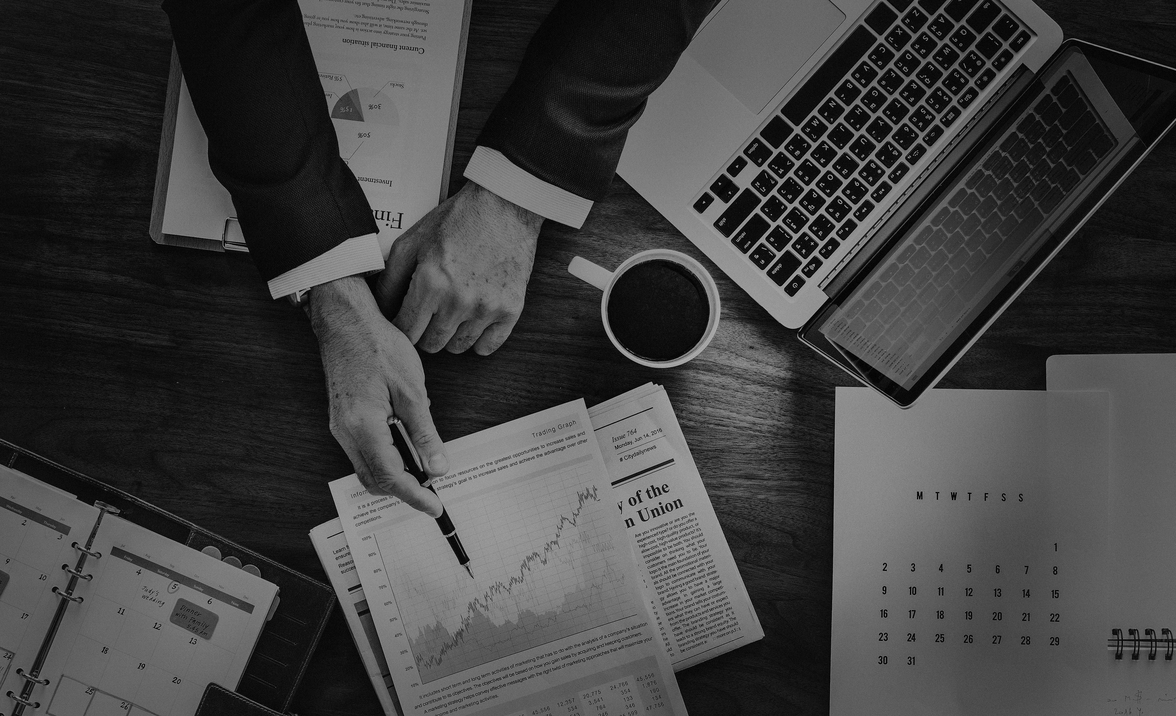 Business 桌布 壁紙 — 俯拍黑白照片，一名商人的雙手在木桌上分析金融圖表和圖形，桌上放有筆記型電腦和咖啡杯。