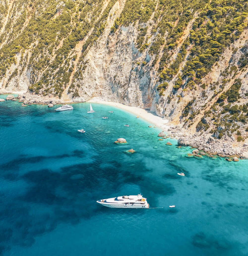 Greek Beach 桌布 壁紙 — 俯瞰一個僻靜的綠松石色海灣，白色遊艇和帆船停泊在一個被陡峭綠色懸崖環抱的月牙形海灘附近。