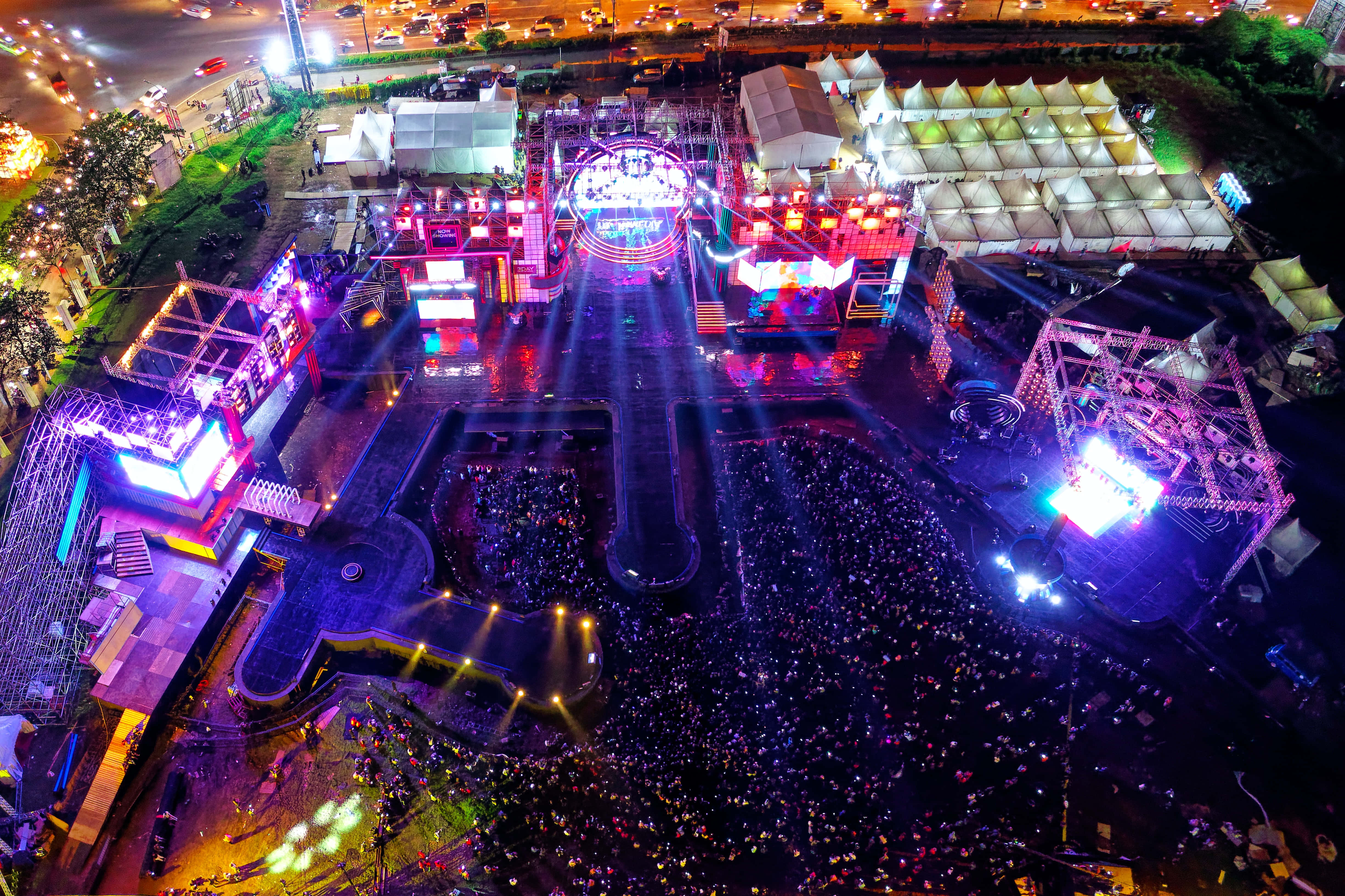 music festival 桌布 壁紙 — 俯瞰一個充滿活力的夜間音樂節，發光的舞台、人群和色彩繽紛的燈光點亮了城市場地。