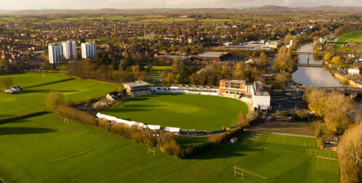 Cricket Ground 桌布 壁紙 — 俯瞰一個充滿活力的綠色橄欖球場，坐落於一個蜿蜒河流和秋樹的廣闊城鎮中。