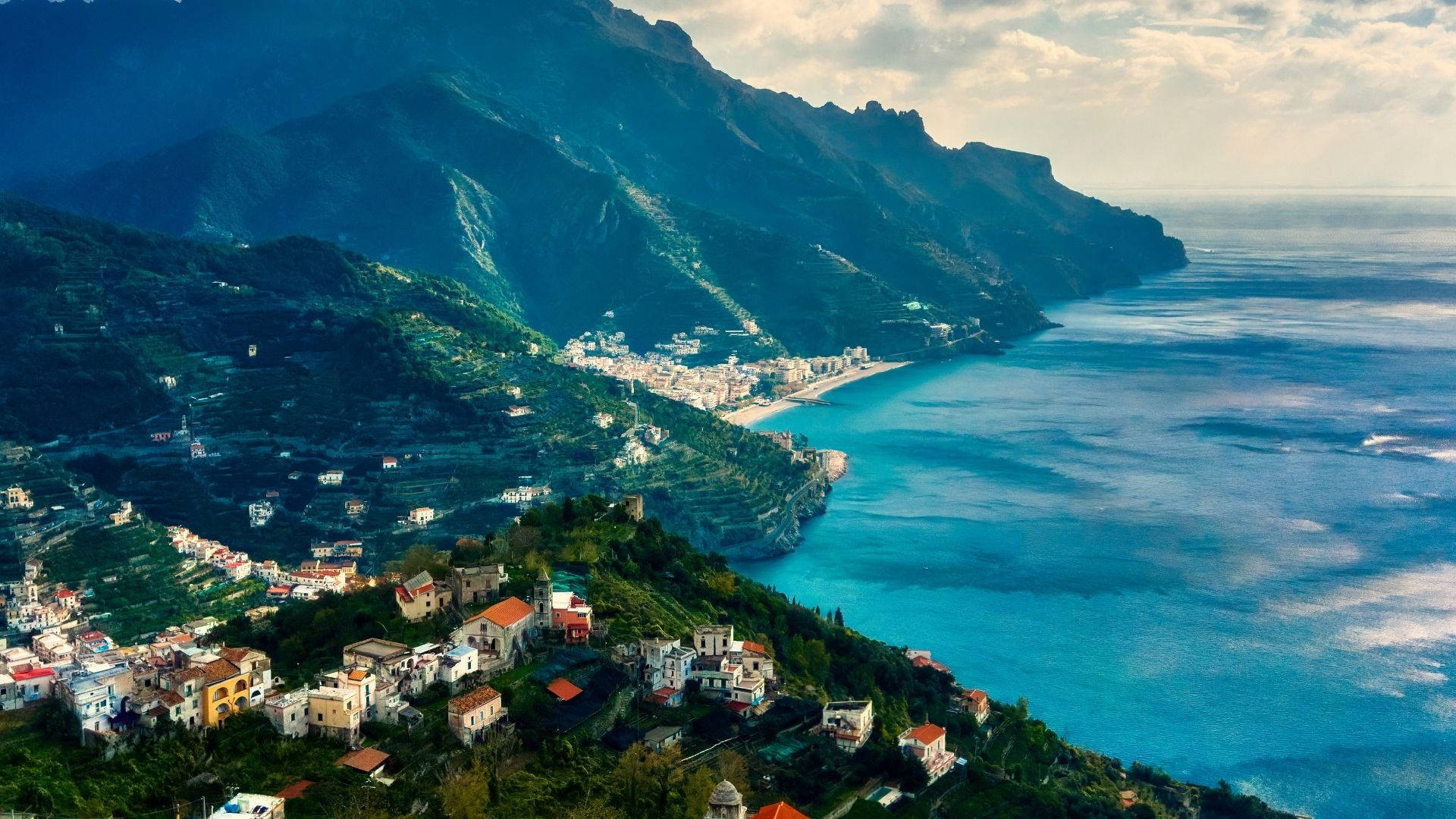 Amalfi Coast 桌布 壁紙 — 俯瞰一個坐落在梯田山丘上的沿海村莊，可欣賞到碧綠的海水和崎嶇的山脈，天空部分多雲。