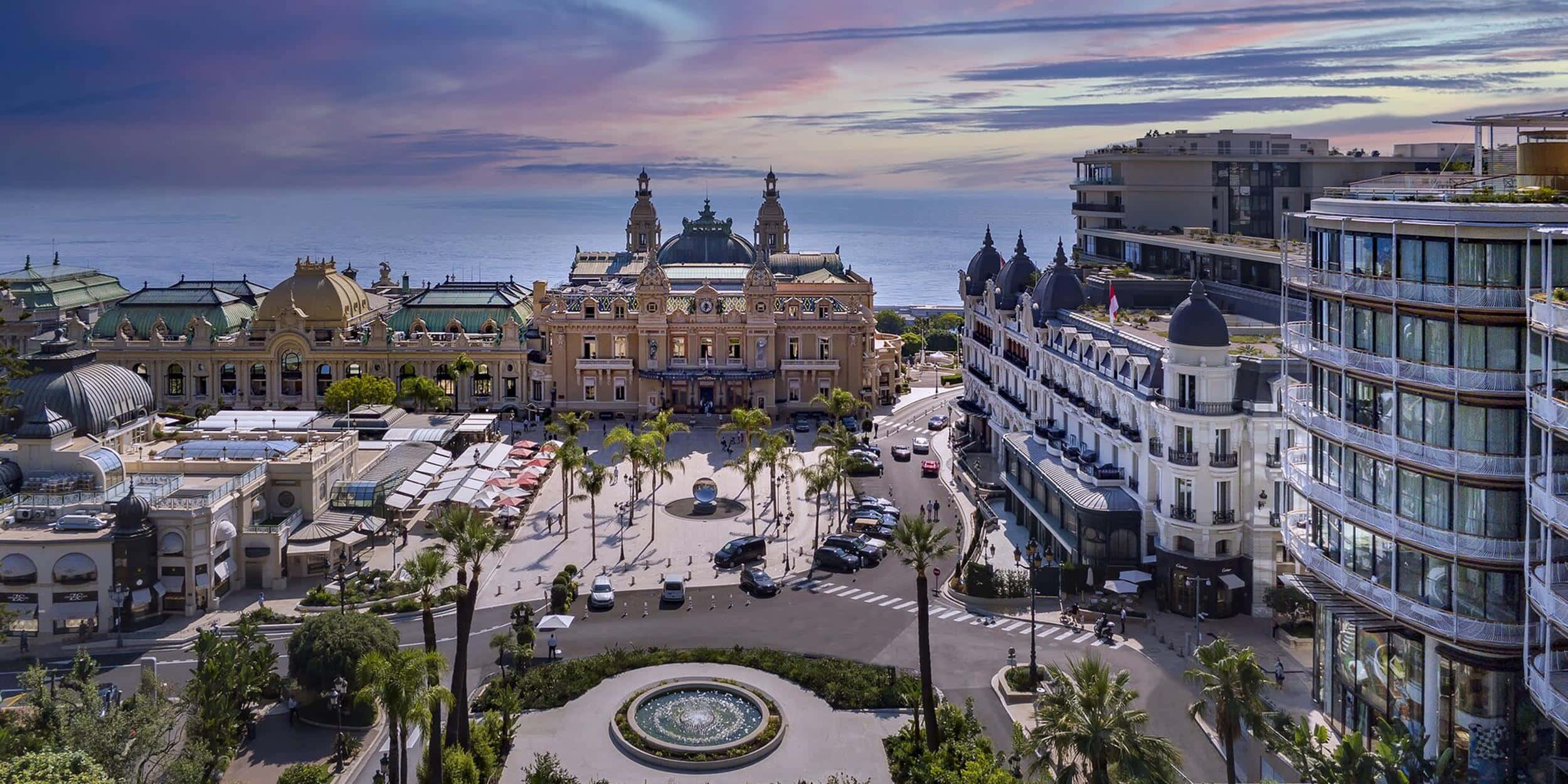 Monaco 桌布 壁紙 — 俯瞰一個宏偉的海岸廣場，擁有華麗的建築、棕櫚樹和一個在日落時俯瞰海洋的噴泉。