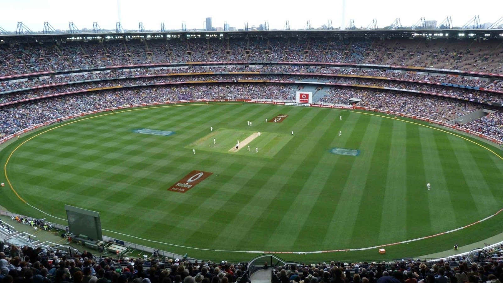 Cricket Ground 桌布 壁紙 — 俯瞰一個擠滿觀眾的板球場，球員在精心劃線的綠色場地上比賽，天空多雲。
