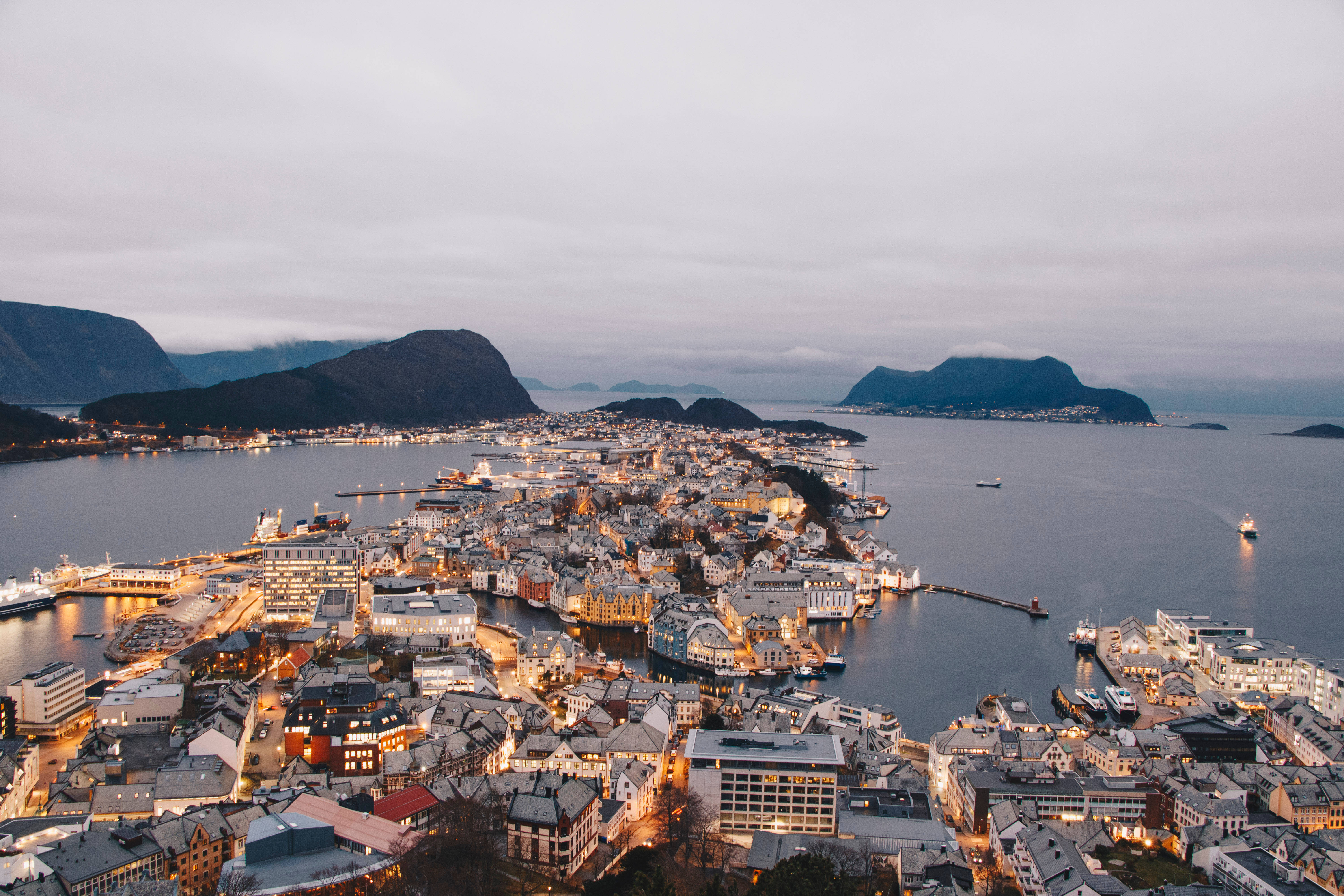 Norway 桌布 壁紙 — 俯瞰一個沿海小鎮，坐落在霧氣繚繞的山巒與平靜峽灣之間，暮色中街燈閃爍。
