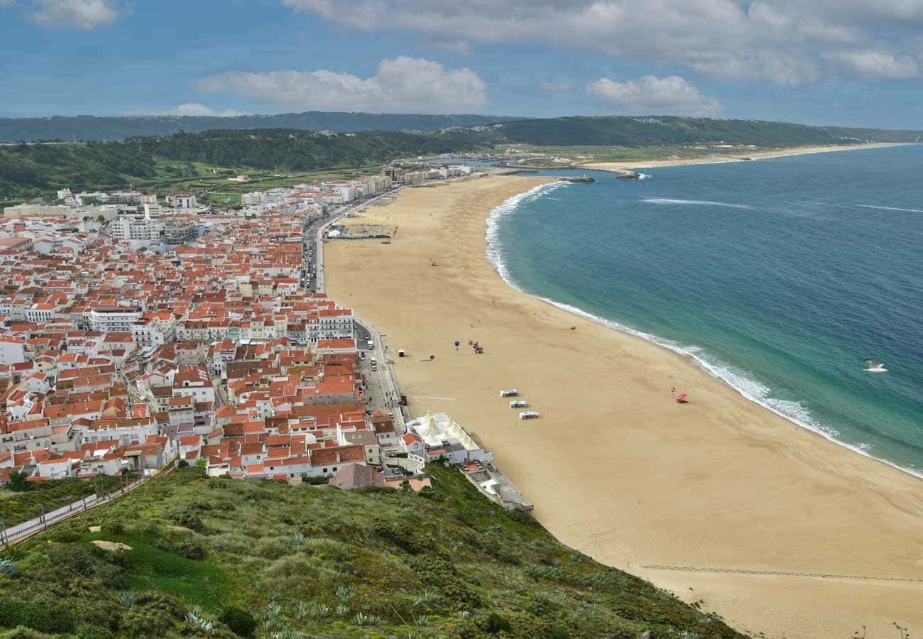 Portuguese Beach 桌布 壁紙 — 俯瞰一個沿海小鎮，紅頂建築依偎著金色沙灘，旁邊是碧綠的海水，天空部分多雲。