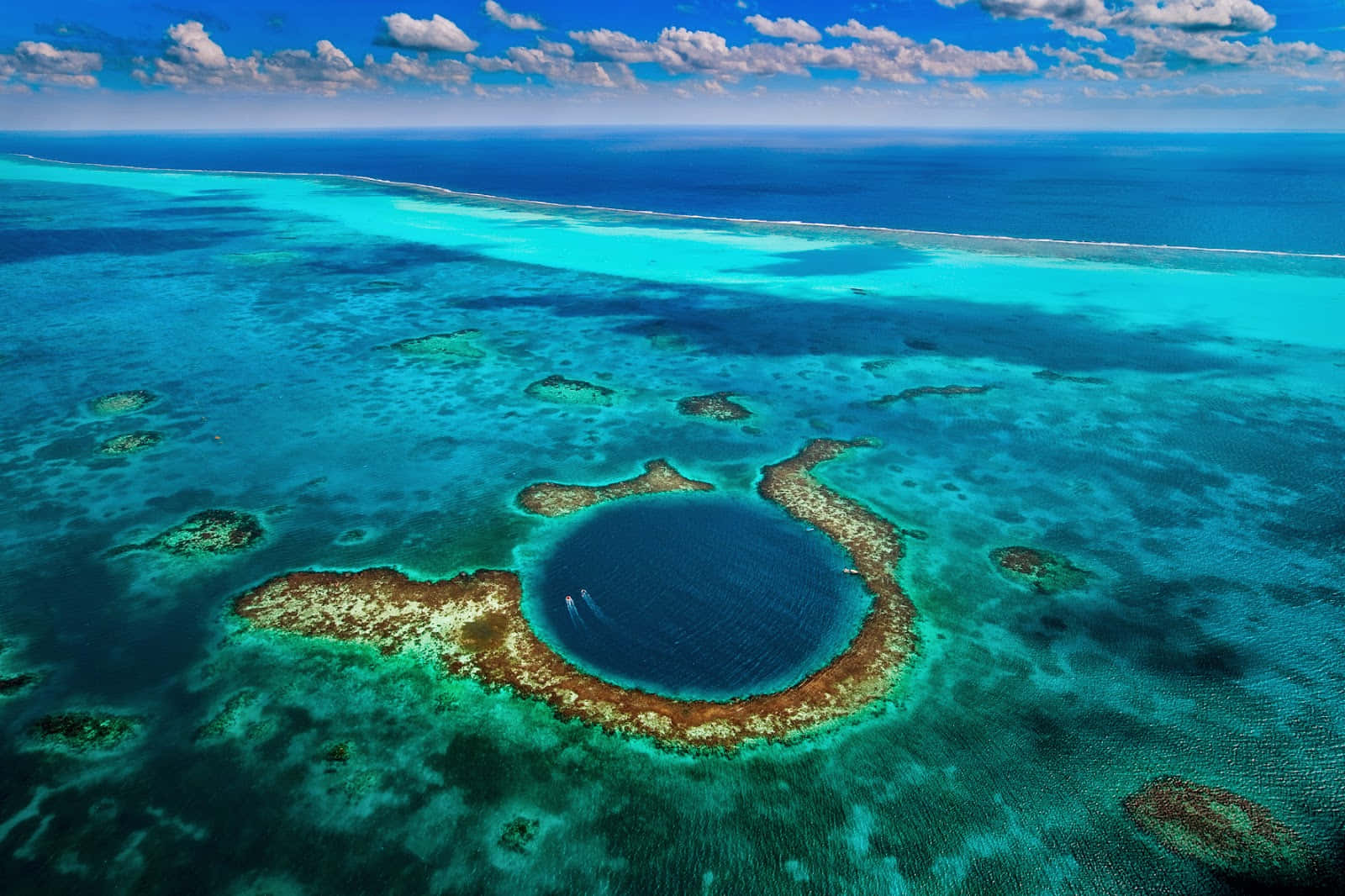 海洋 桌布 壁紙 — 俯瞰一個由珊瑚礁和碧綠海水環繞的迷人圓形藍色潟湖，在明亮的藍天和散落的雲朵下。