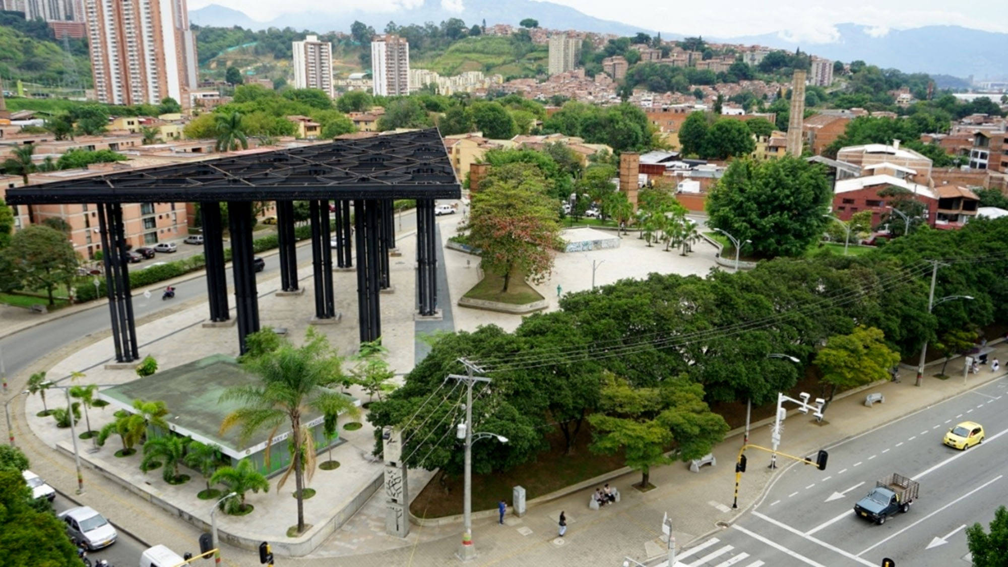 Medellin 桌布 壁紙 — 俯瞰一個繁忙的城市公園中的現代黑色鋼結構，周圍環繞著綠樹和城市建築，天空多雲。