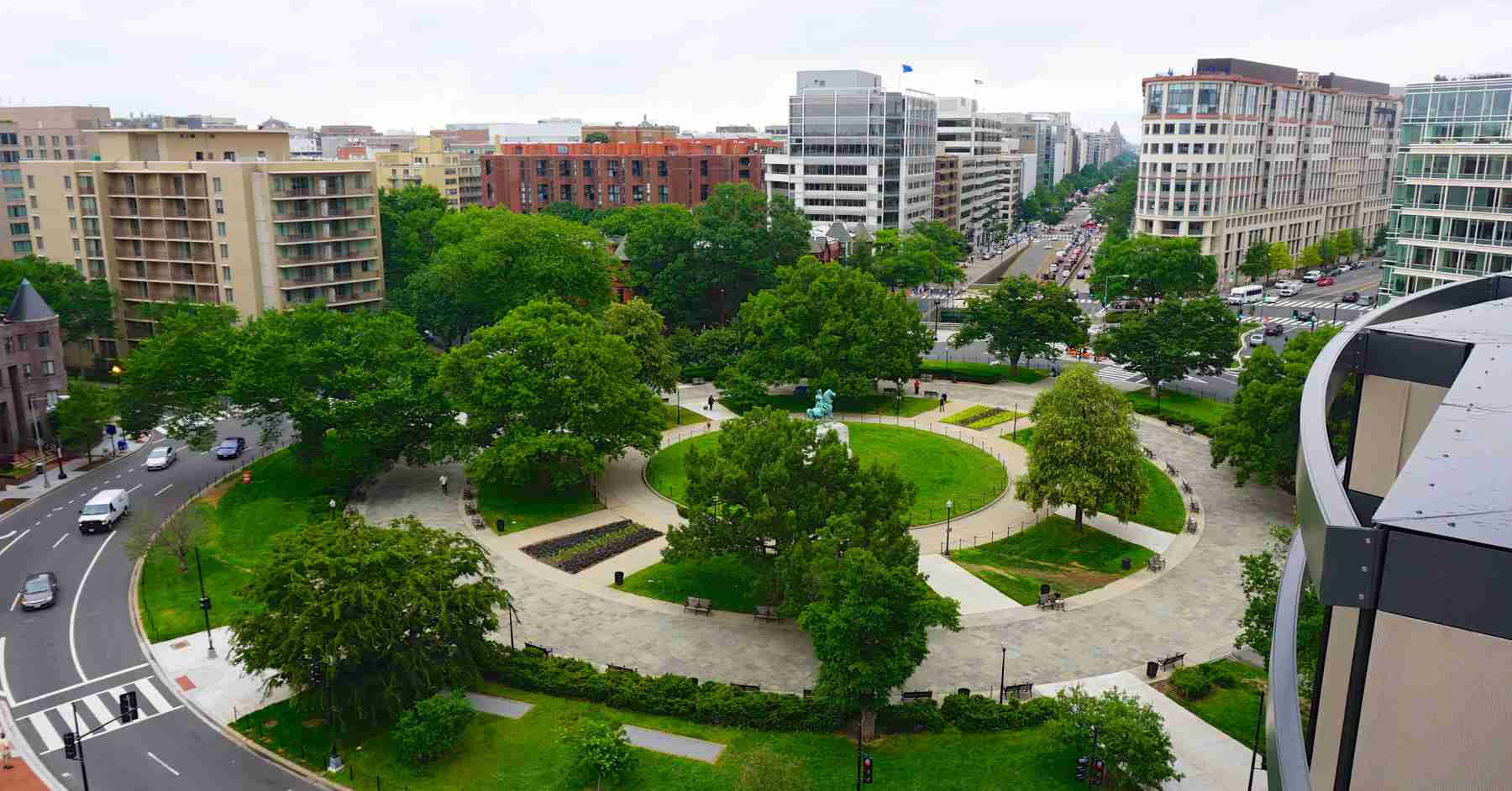 學校 桌布 壁紙 — 俯瞰一個鬱鬱蔥蔥的綠色城市公園，有蜿蜒小徑和成熟樹木，周圍是現代和住宅建築，天空多雲。