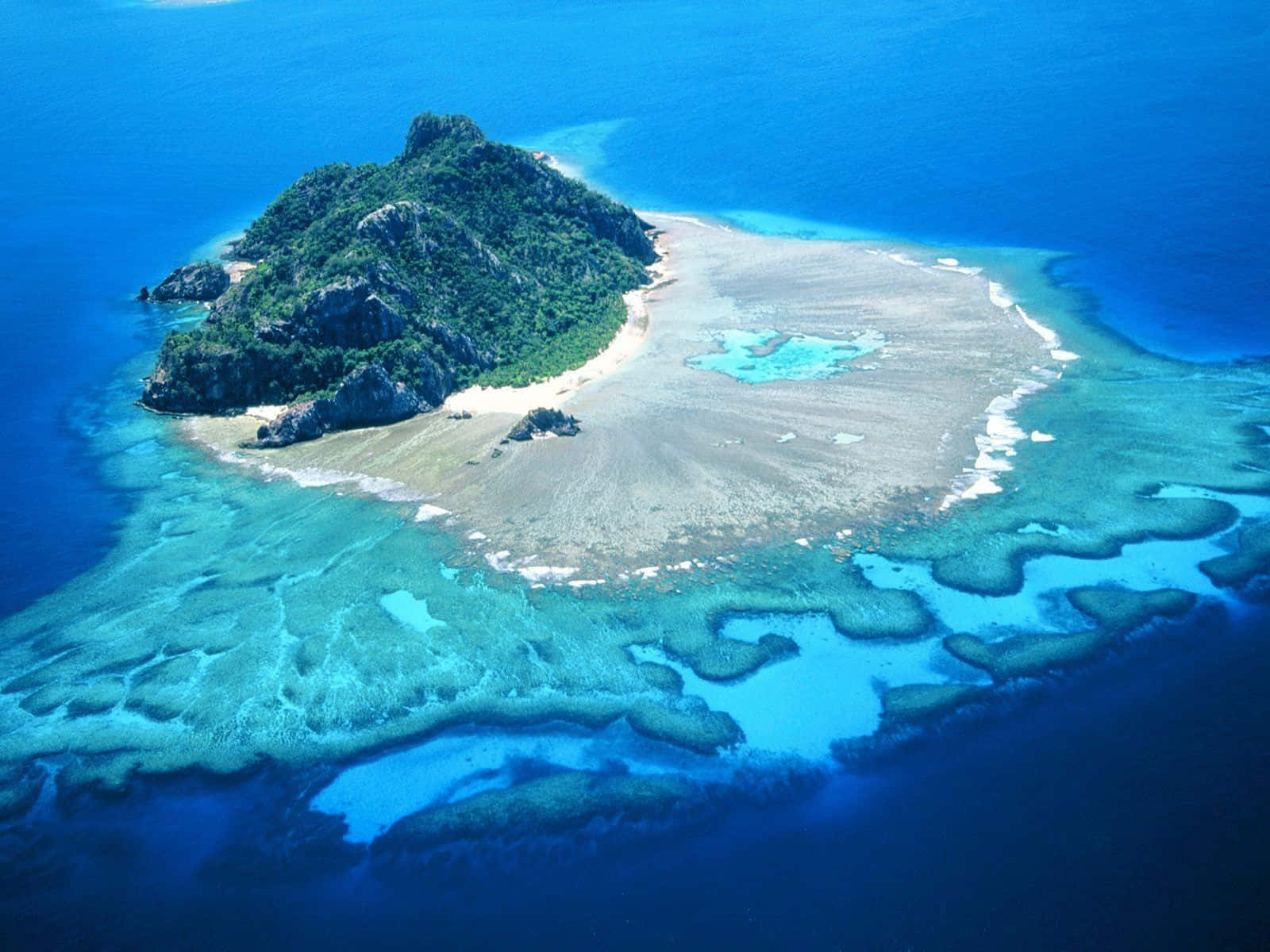 島嶼 桌布 壁紙 — 俯瞰一個鬱鬱蔥蔥的綠色島嶼，周圍環繞著充滿活力的綠松石珊瑚礁和深藍色海洋。