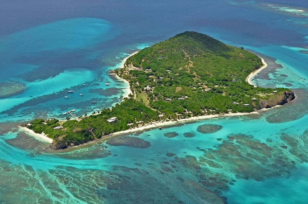 Caribbean Island 桌布 壁紙 — 俯瞰一個鬱鬱蔥蔥的綠色島嶼，周圍環繞著充滿活力的碧綠海水和珊瑚礁，船隻停泊在岸邊附近。