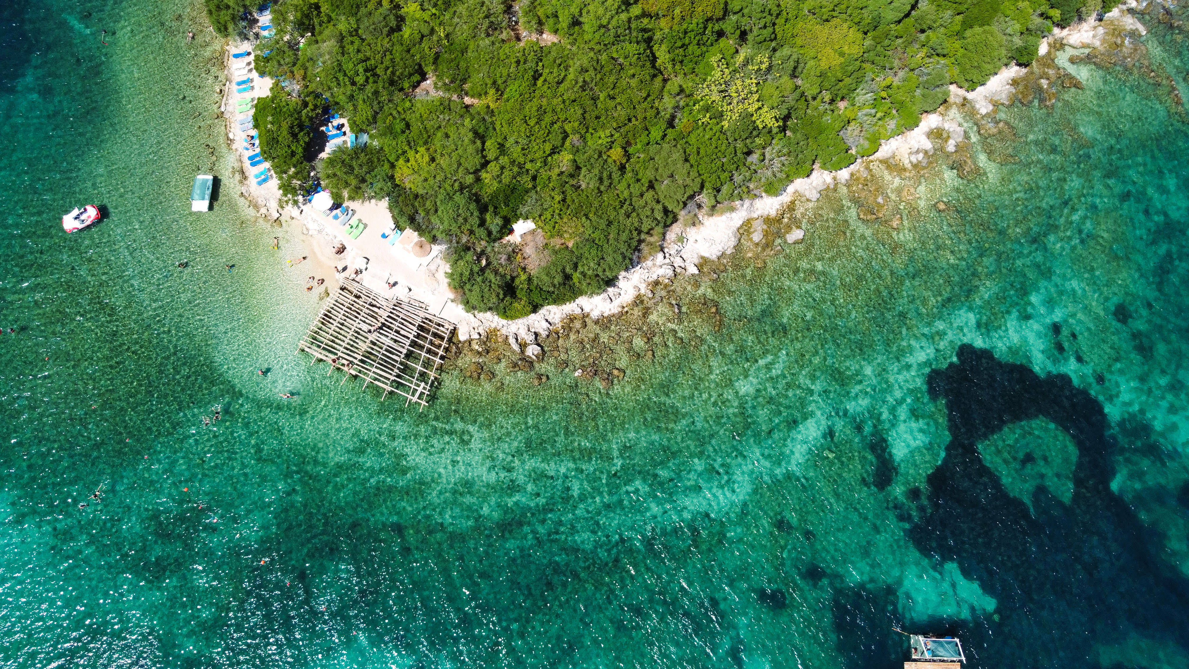Albania 桌布 壁紙 — 俯瞰一個鬱鬱蔥蔥的綠色島嶼，擁有碧綠的海水和沙灘，沙灘上排列著藍色遮陽傘和曬太陽的人。