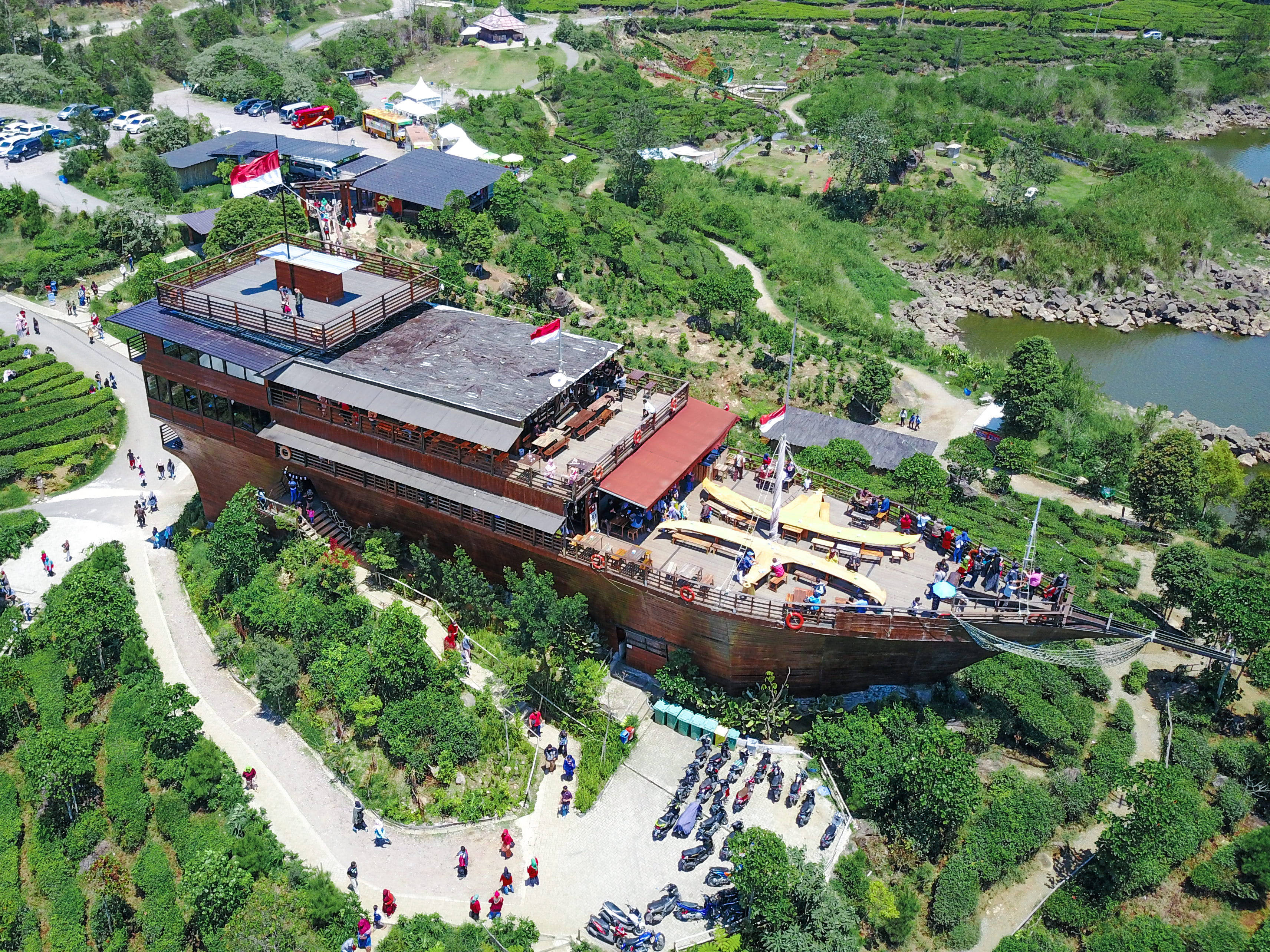 bandung 桌布 壁紙 — 俯瞰一座巨大的木造船形建築，坐落在鬱鬱蔥蔥的綠色茶園梯田中，遊客正在探索其甲板和周圍的小徑。