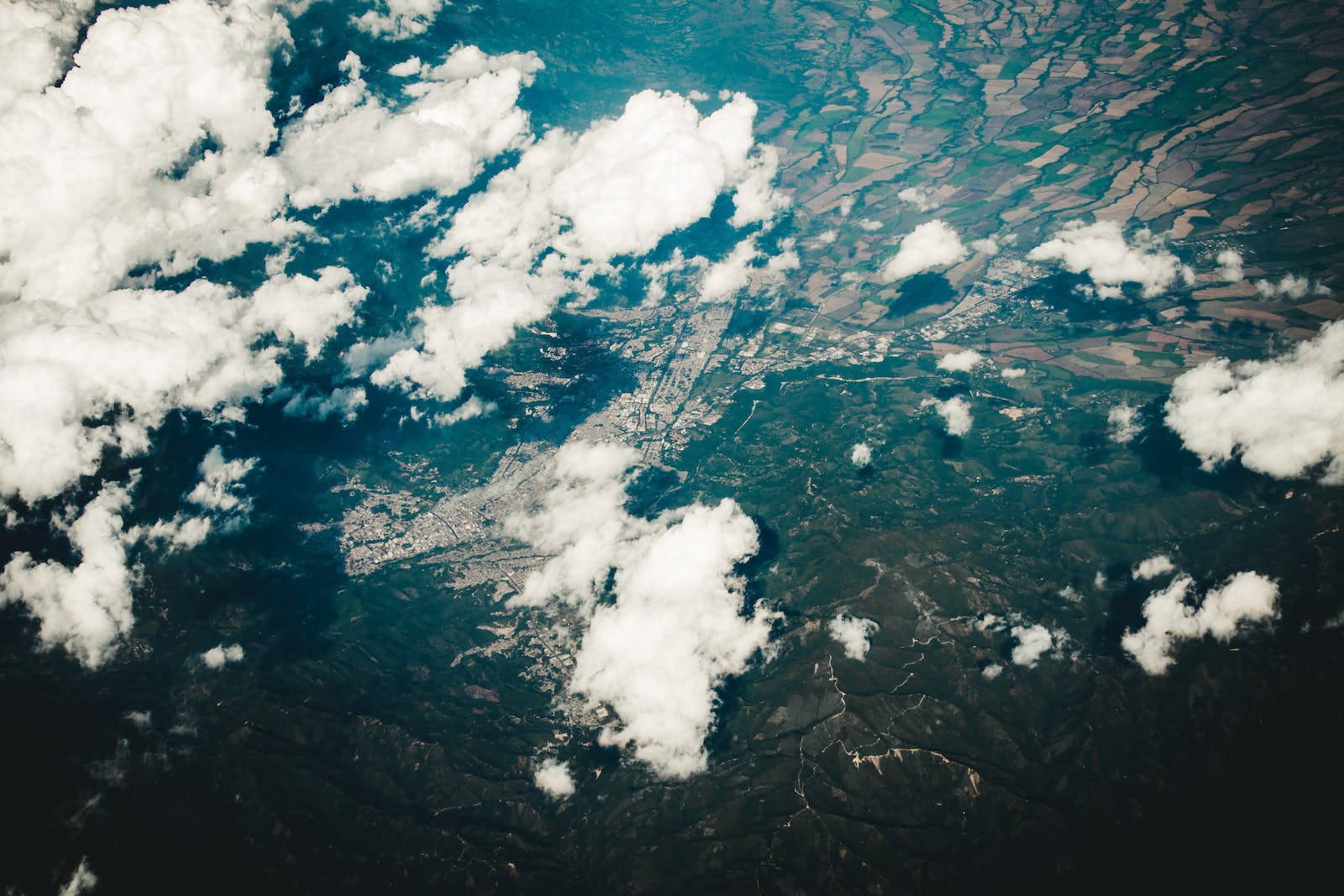 地形 桌布 壁紙 — 俯瞰一座廣闊城市，坐落於翠綠山丘與農田之間，部分被蓬鬆白雲遮蔽。