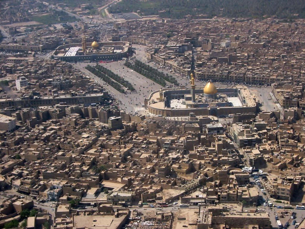 Karbala 桌布 壁紙 — 俯瞰一座廣闊的沙漠城市，主體是兩個金色圓頂宗教建築群，周圍環繞著密集的傳統建築。