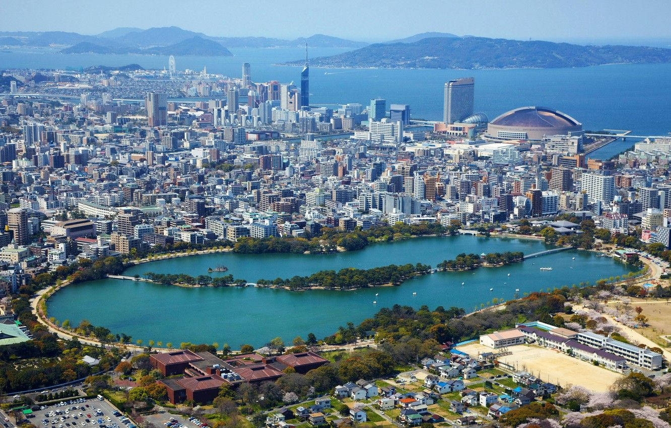 Fukuoka 桌布 壁紙 — 俯瞰一座廣闊的海濱城市，碧綠的湖泊被綠植和現代摩天大樓環繞，天空湛藍清澈。