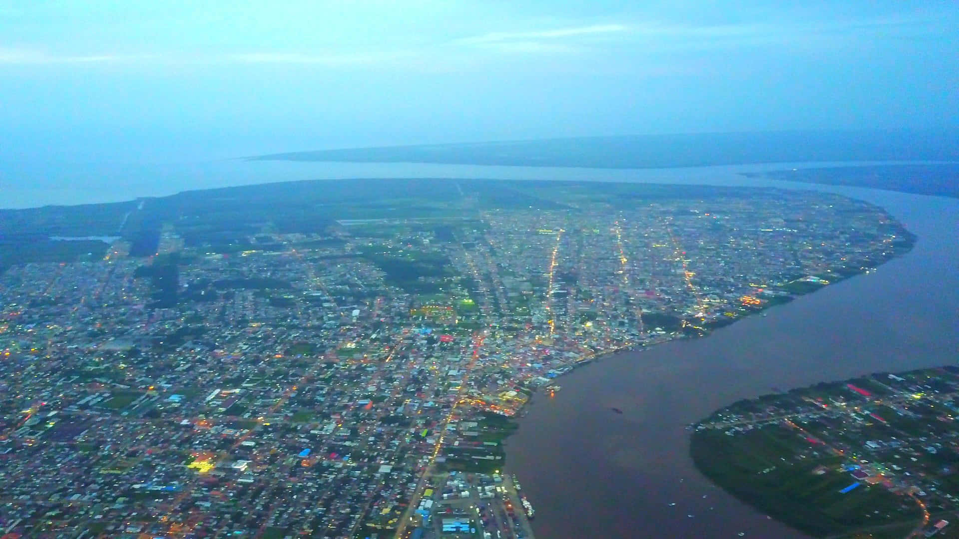 World Travel 桌布 壁紙 — 俯瞰一座沿著寬闊河流而建的廣闊城市，在暮色中閃爍著街燈和建築物的燈光。