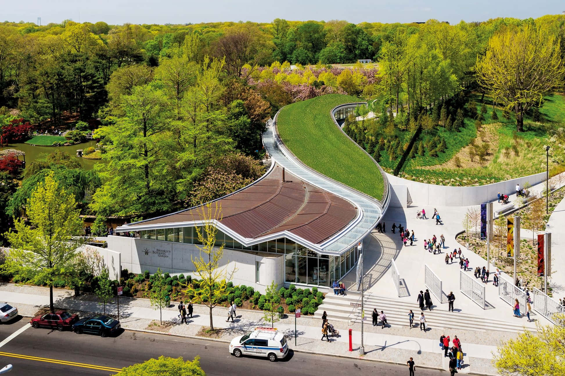 Botanical Garden 桌布 壁紙 — 俯瞰一座現代玻璃與混凝土建築，坐落於鬱鬱蔥蔥的公園中，蜿蜒小徑上有人們漫步。
