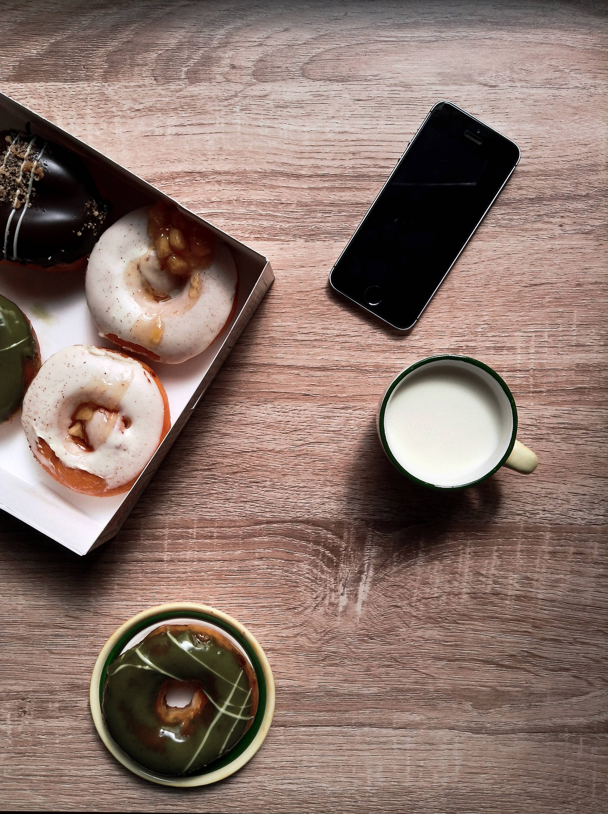 food iPhone 桌布 壁紙 — 俯瞰一盒綜合甜甜圈，旁邊有一杯牛奶和一支智慧型手機，放在木桌上。