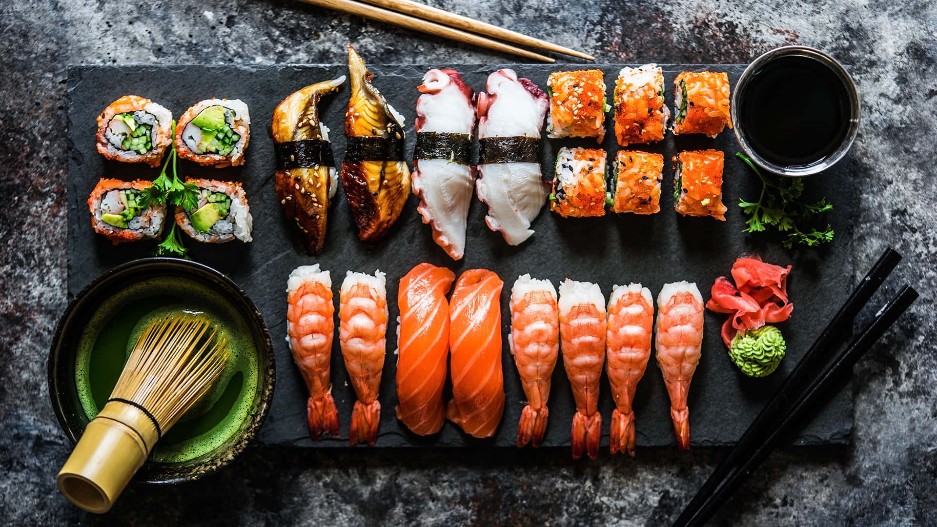 Sushi 桌布 壁紙 — 俯瞰各種壽司卷、刺身和裝飾，整齊排列在深色石板盤上，搭配醬油和芥末。