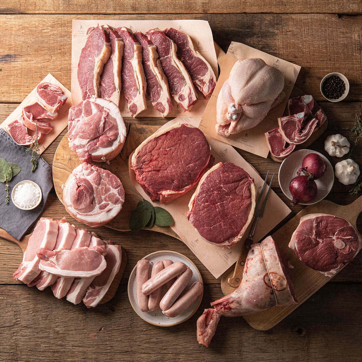 Red Meat 桌布 壁紙 — 俯瞰各類生肉，包括牛排、豬排、香腸及整塊切肉，排列在木板上，搭配大蒜、洋蔥及香草，置於鄉村風木桌上。
