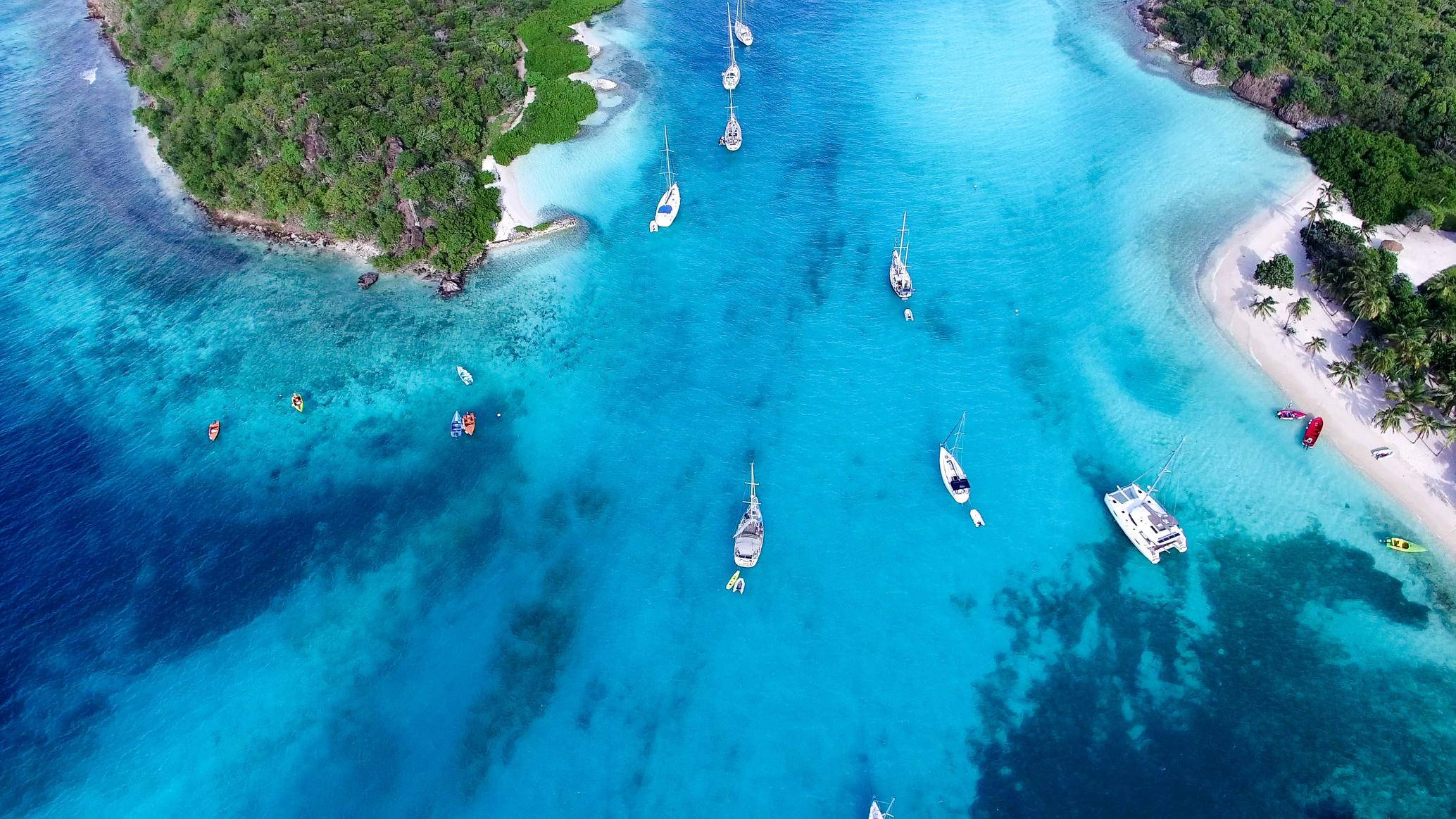 St Vincent And The Grenadines 桌布 壁紙 — 俯瞰圖：翠綠島嶼環繞的白色沙灘附近，一艘艘帆船和獨木舟點綴在碧綠的潟湖上。