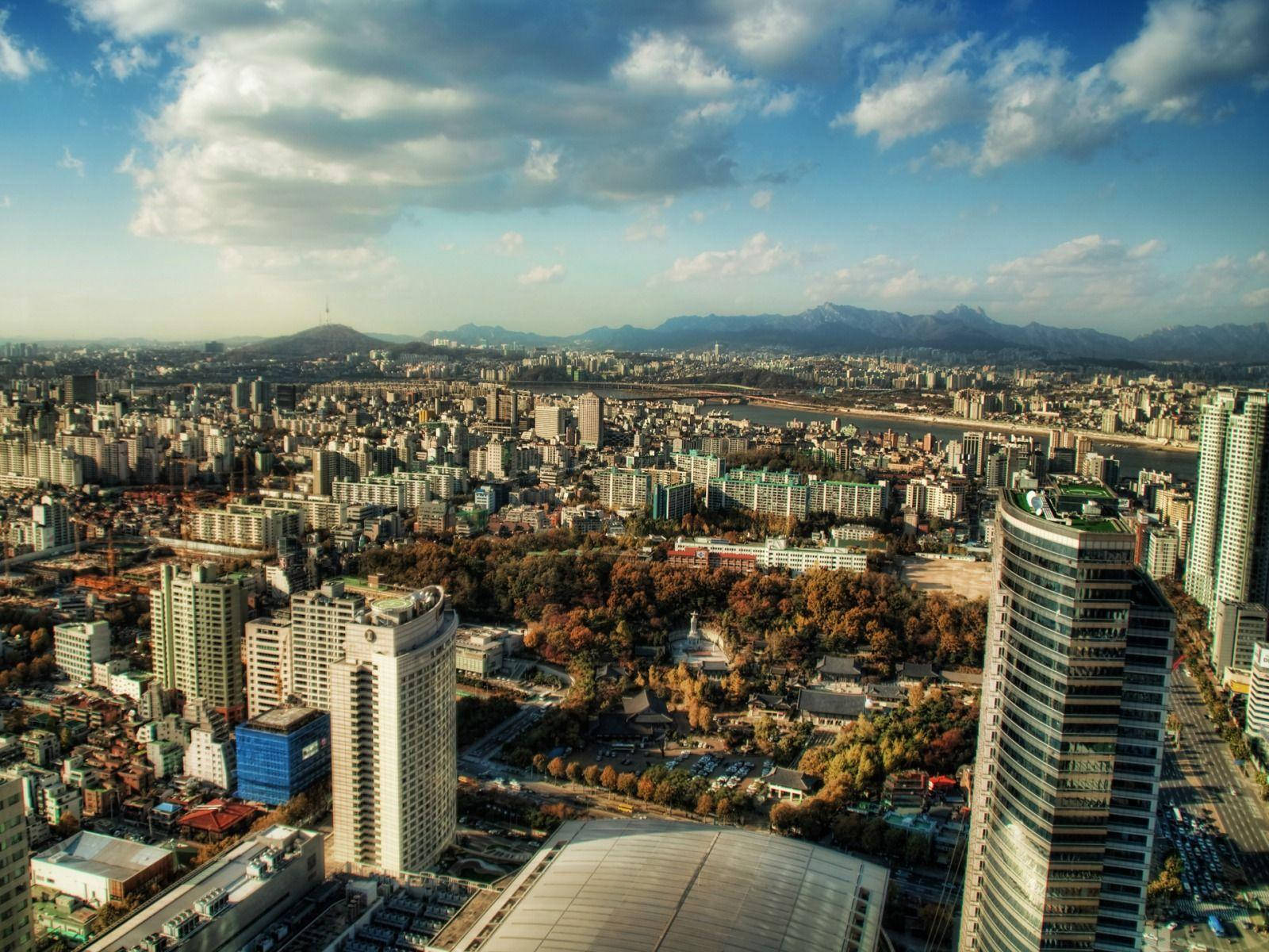 南韓 桌布 壁紙 — 俯瞰廣闊的城市景觀，秋樹與遠山在部分多雲的天空下。