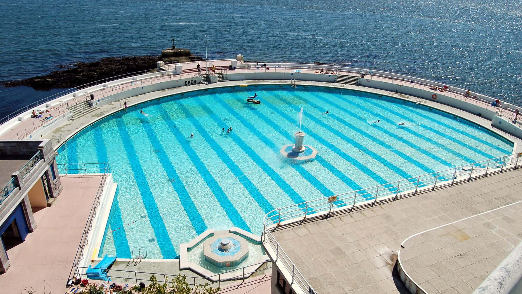 Beach Swimming Pool 桌布 壁紙 — 俯瞰彎曲的海濱泳池，泳客在條紋藍色海水中游泳，旁邊是岩石海岸線。