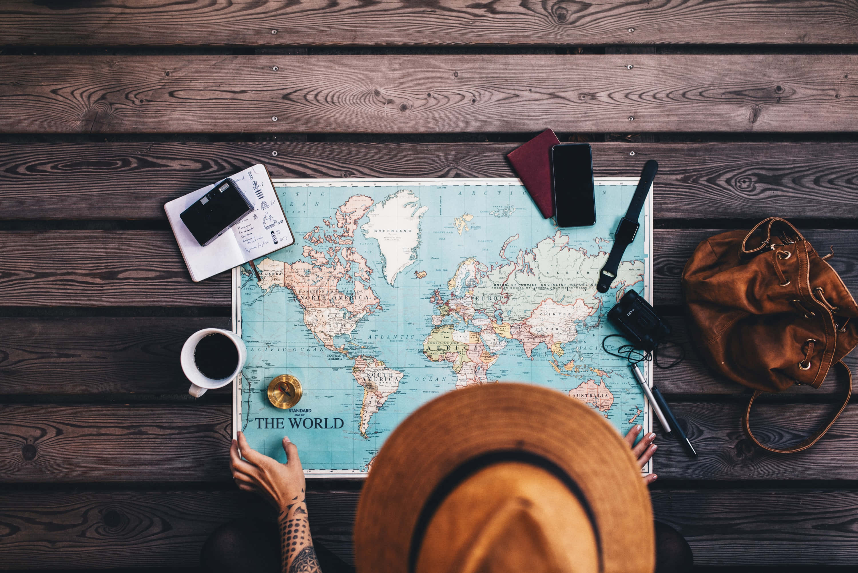 Travel Desktop 桌布 壁紙 — 俯瞰視角，一位戴帽子的旅行者在木桌上，被旅行裝備包圍，正在世界地圖上規劃旅程。