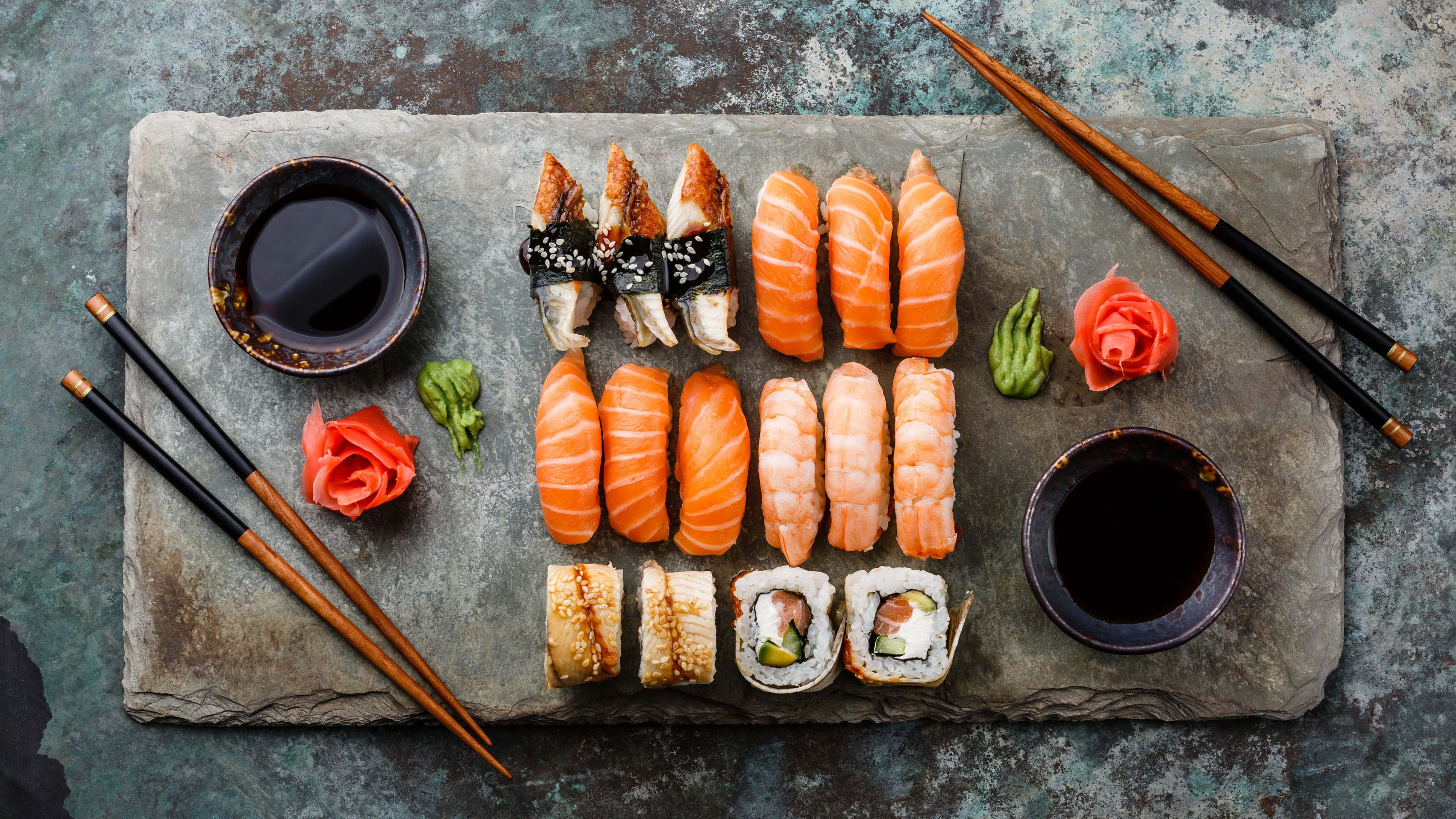 Sushi 桌布 壁紙 — 俯瞰視角，各種壽司卷和握壽司整齊排列在質樸的石板上，搭配醬油和筷子。