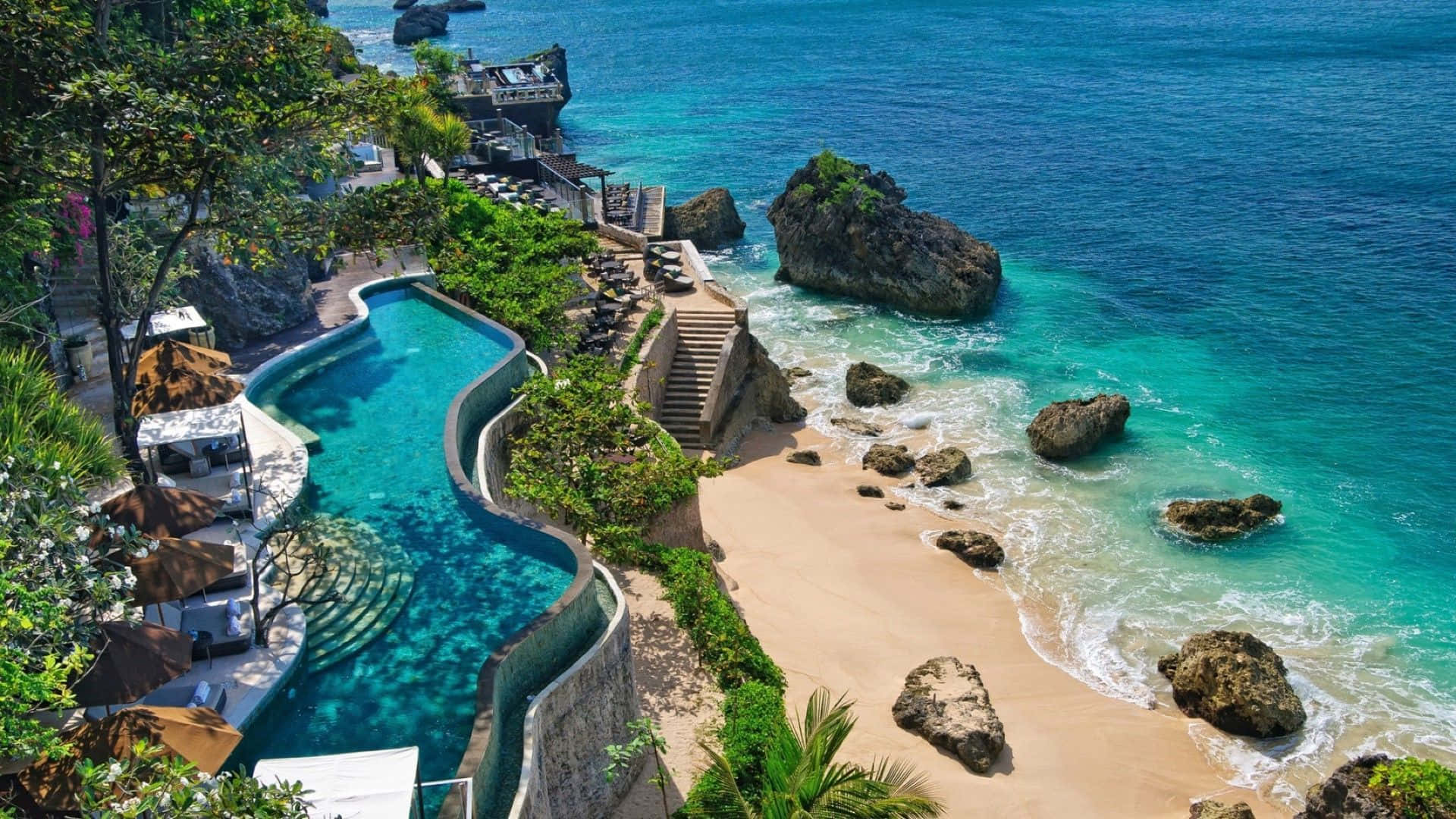Bali 桌布 壁紙 — 俯瞰豪華無邊際泳池蜿蜒於熱帶海灘，伴隨碧綠海洋與岩石露頭的景觀。