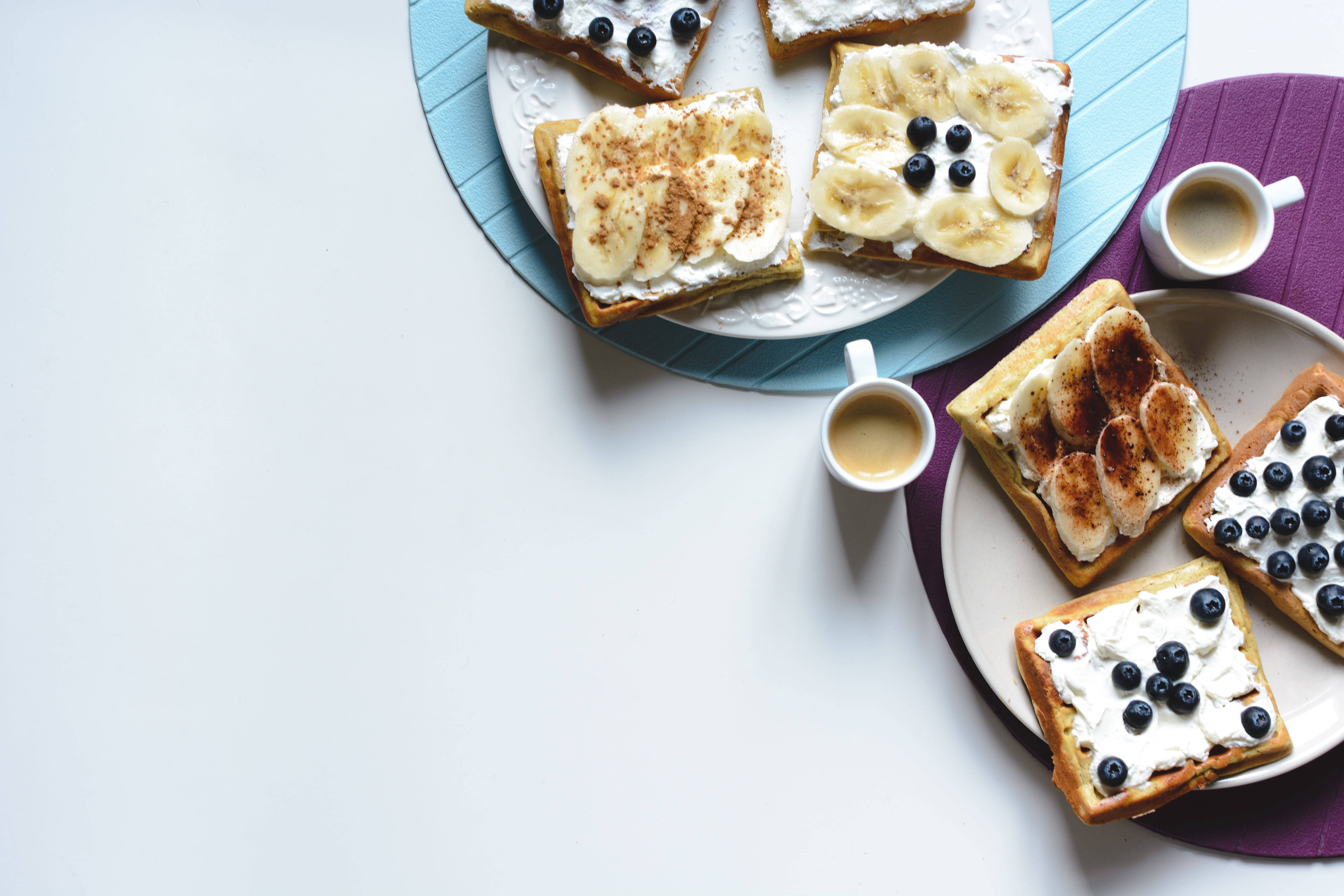 Waffle 桌布 壁紙 — 俯瞰金黃色鬆餅，上面有鮮奶油、香蕉和藍莓，搭配咖啡，放在彩色盤子上。