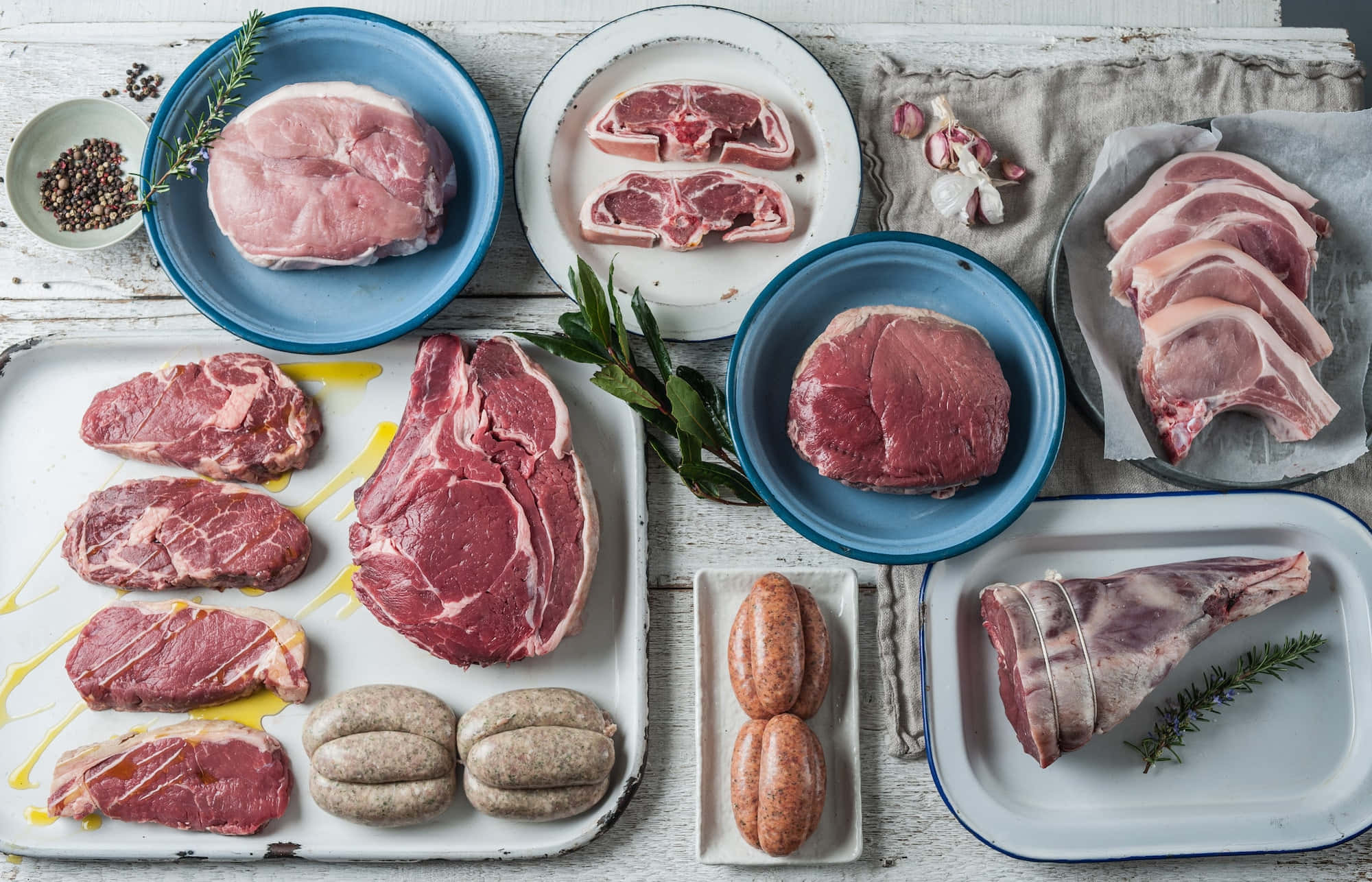 Red Meat 桌布 壁紙 — 俯瞰鏡頭下，各種生肉包括豬排、牛排、香腸和羊腿，放在盤子上，配上香草和油，置於鄉村風格的木質表面上。