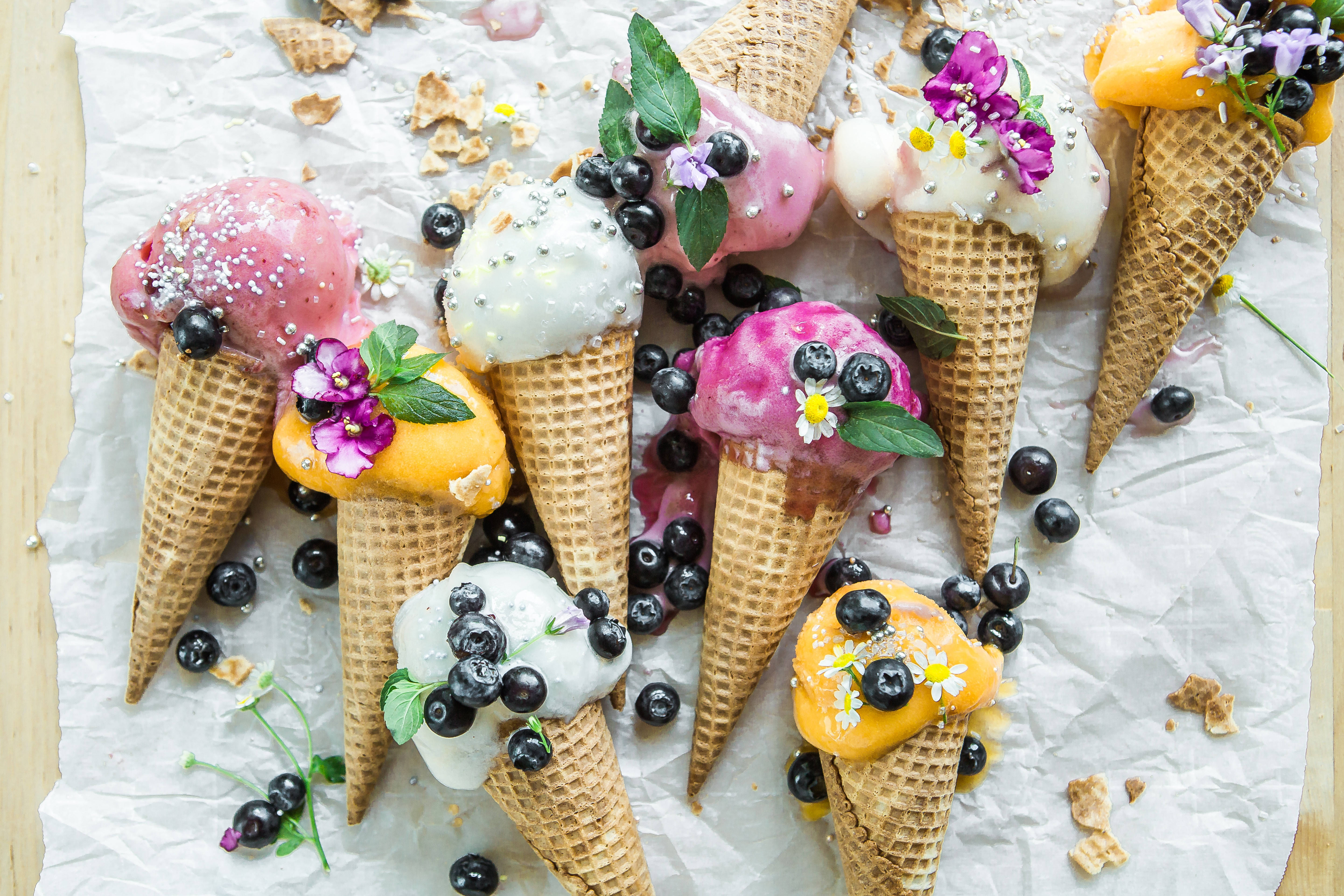 Ice Cream 桌布 壁紙 — 俯瞰鏡頭下，色彩繽紛的冰淇淋甜筒上綴有莓果、花朵和糖粒，放在皺巴巴的烘焙紙上。