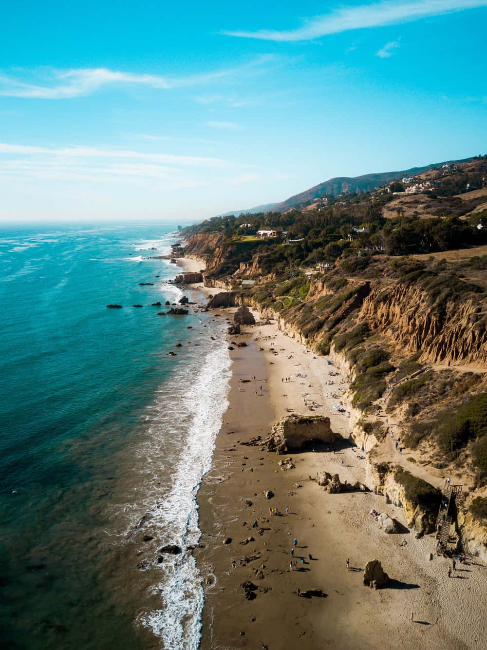 Malibu 桌布 壁紙 — 俯瞰陽光普照的海岸沙灘，碧綠的海浪拍打著金色沙灘，旁邊是陡峭的懸崖，上面散落著房屋。