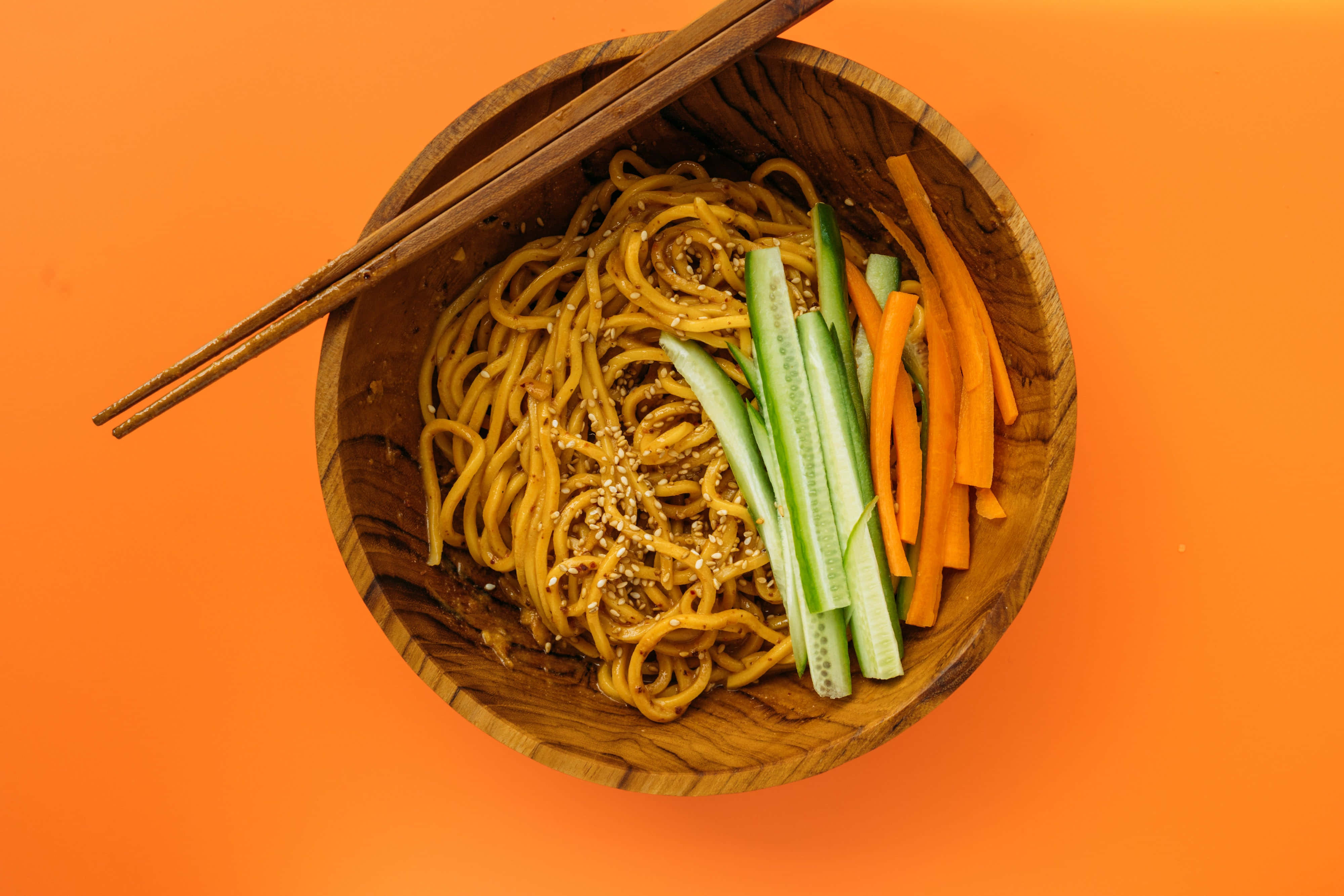Noodles 桌布 壁紙 — 俯視一個木碗，裡面裝滿了芝麻麵條和切片小黃瓜及紅蘿蔔，背景是鮮豔的橙色。