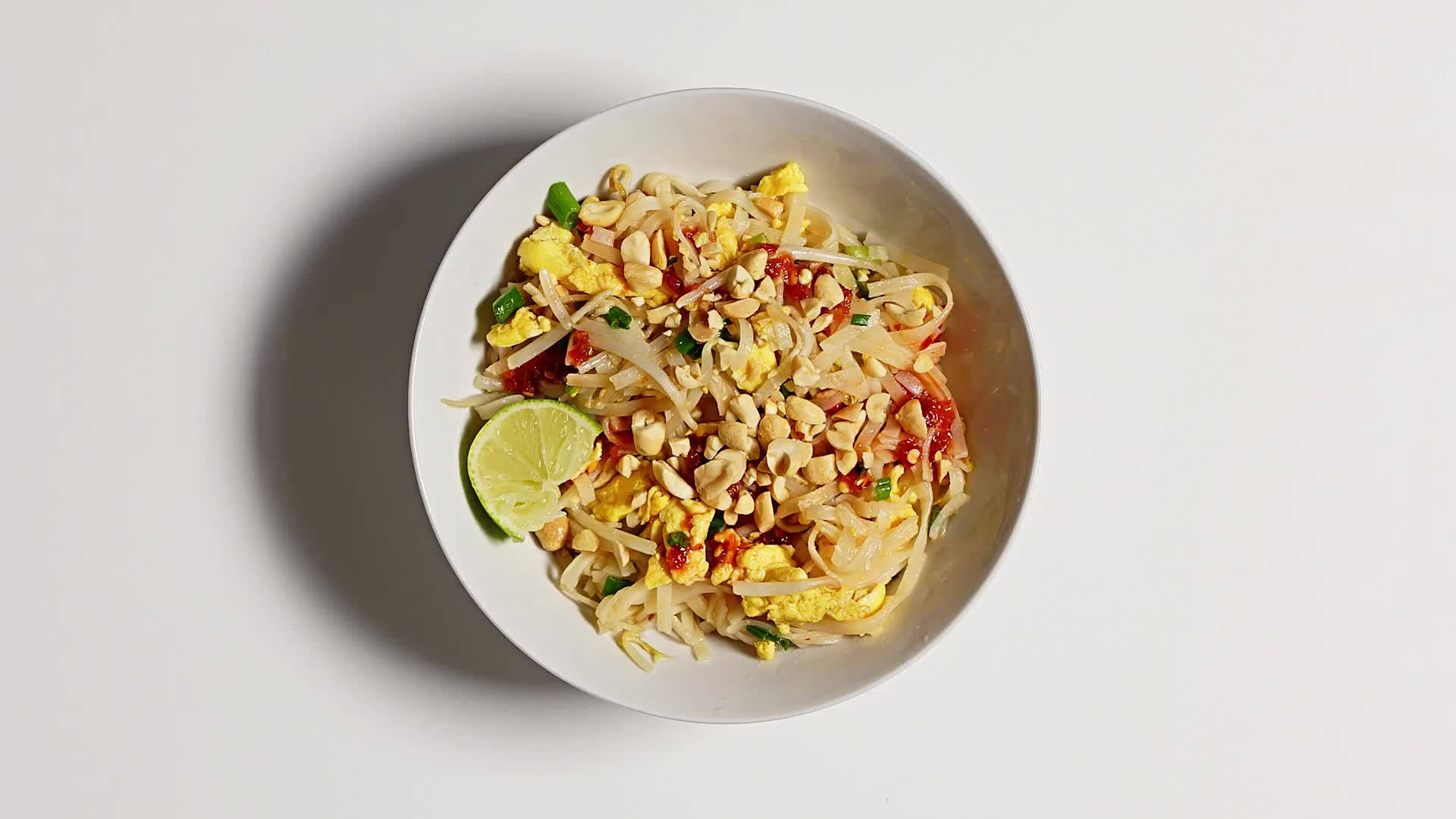 thai 桌布 壁紙 — 俯視一個白色碗，裡面裝滿了色彩鮮豔的泰式炒河粉，上面撒有花生、青檸和辣椒。