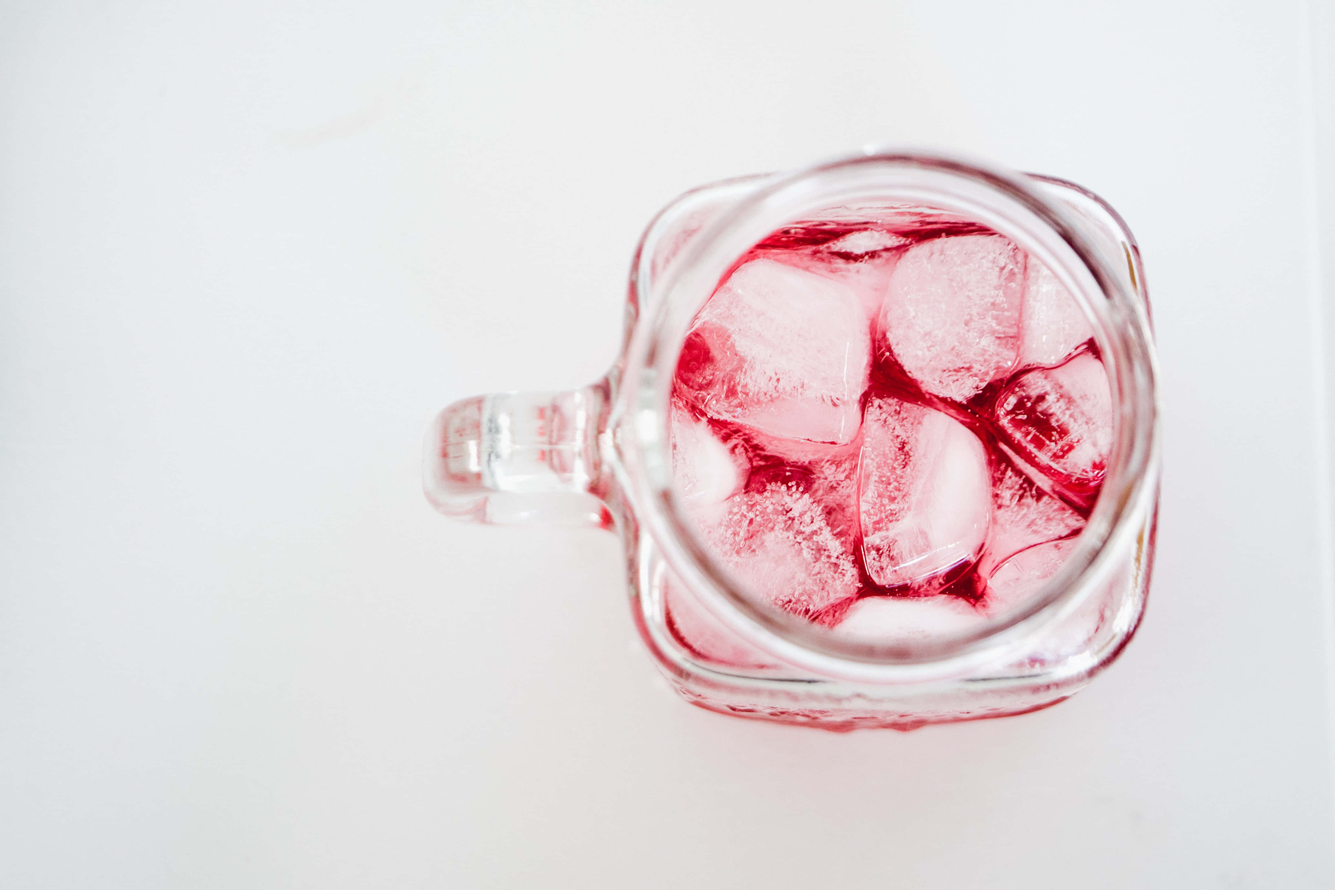 Summer Drinks 桌布 壁紙 — 俯視一個透明玻璃杯，裝有粉紅色液體和正在融化的冰塊，放置在白色表面上。
