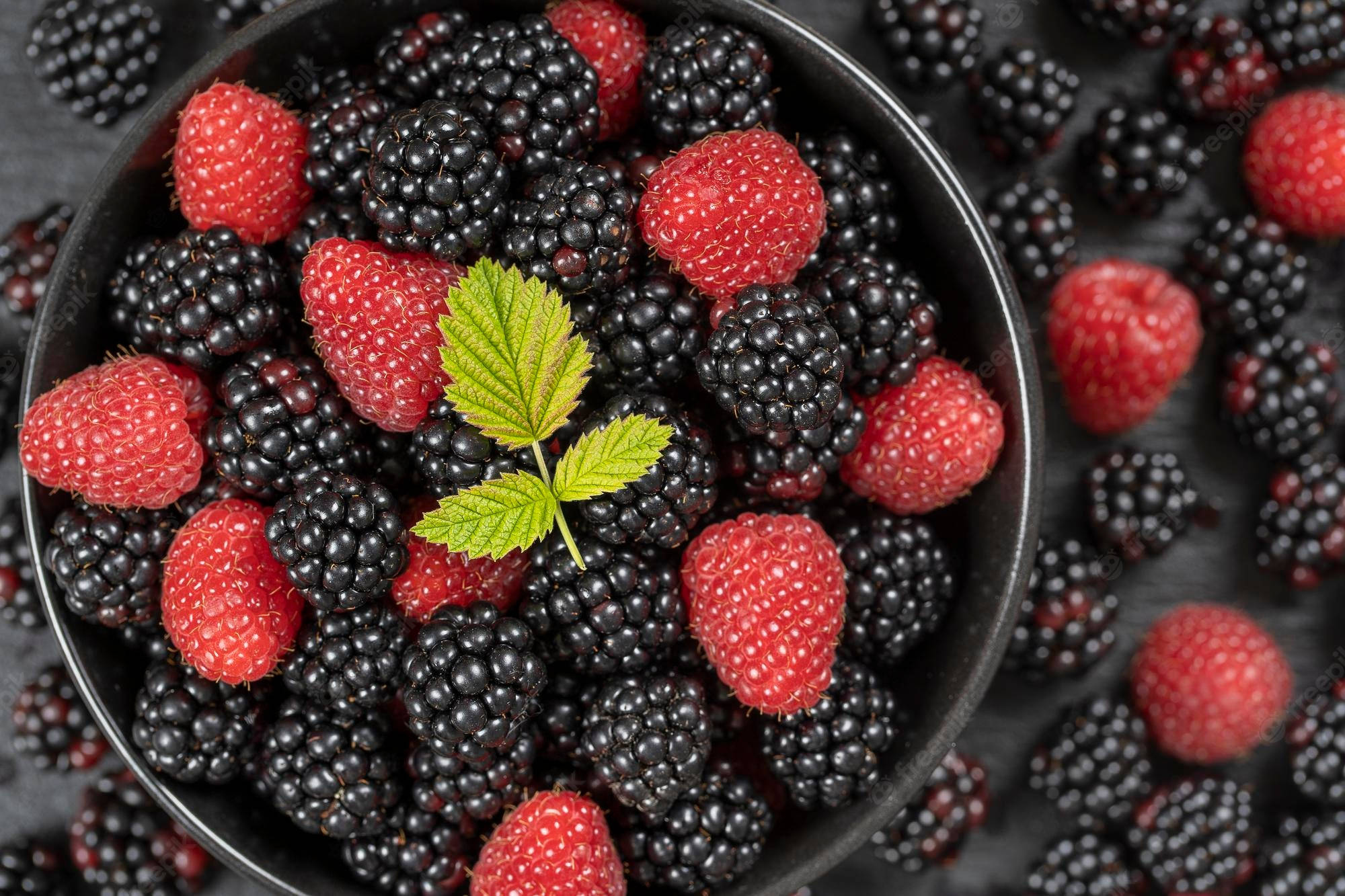 Berry 桌布 壁紙 — 俯視一個黑色碗，裡面裝滿鮮紅色的覆盆子和光澤的黑莓，並以一片綠葉點綴。