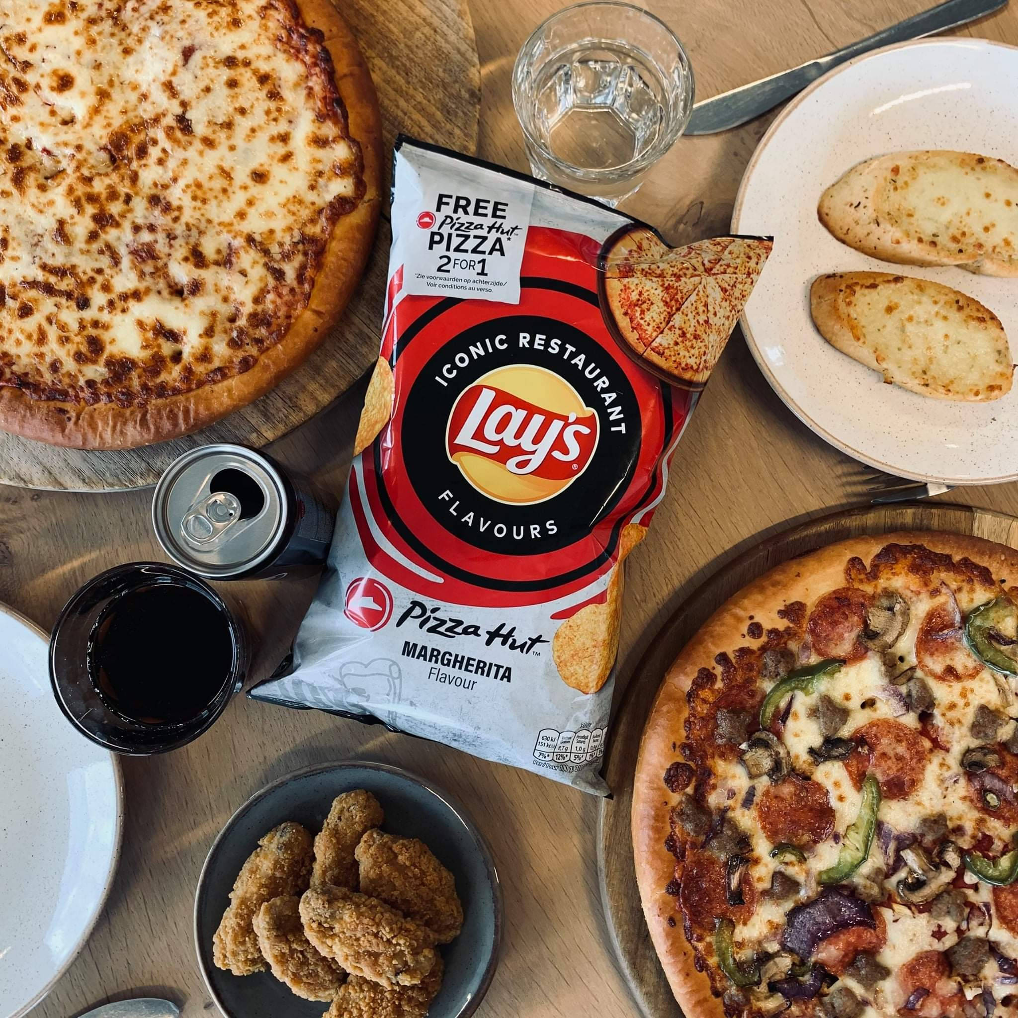 Hut 桌布 壁紙 — 俯視一張木桌上放有兩個披薩、炸雞、麵包棒和一袋Lay's Pizza Hut Margherita口味洋芋片。