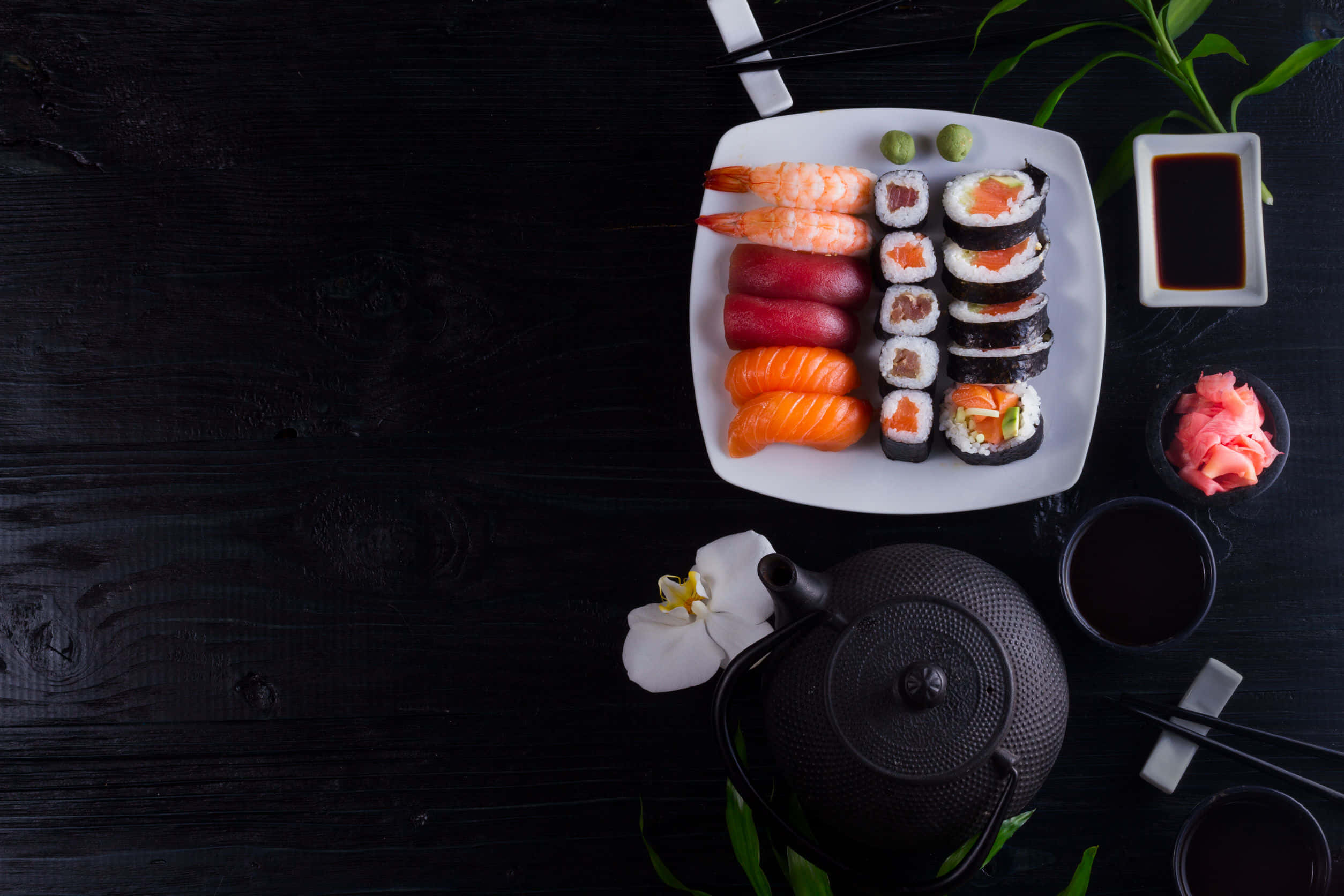 Sushi 桌布 壁紙 — 俯視圖，一個白色盤子上放滿各式壽司和生魚片，整齊地排列在深色木桌上，配有茶具和裝飾。