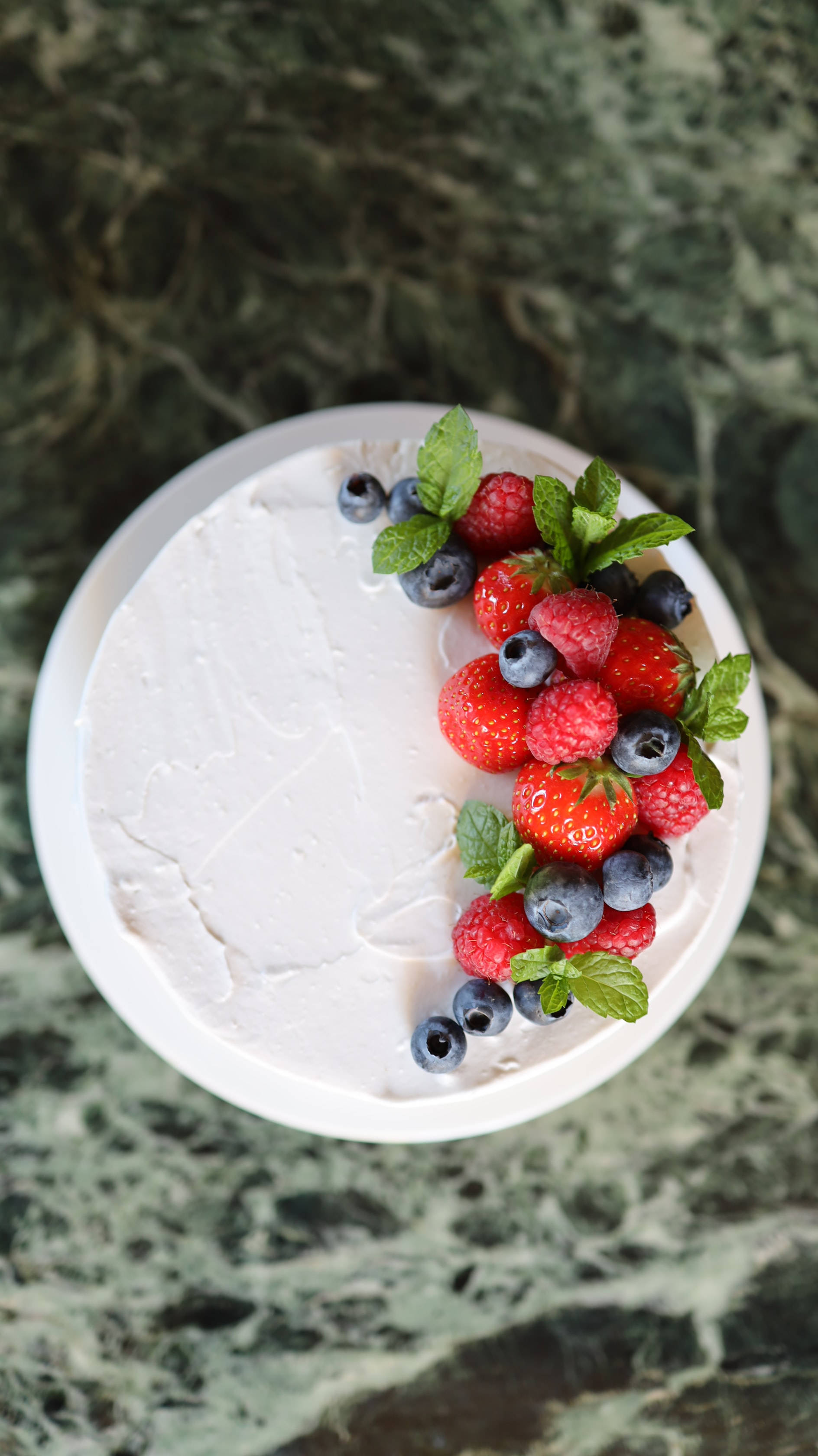 Cake 桌布 壁紙 — 俯視圖：一個白色糖霜蛋糕，上面點綴著新鮮草莓、覆盆子、藍莓和薄荷葉，置於綠色大理石表面上。