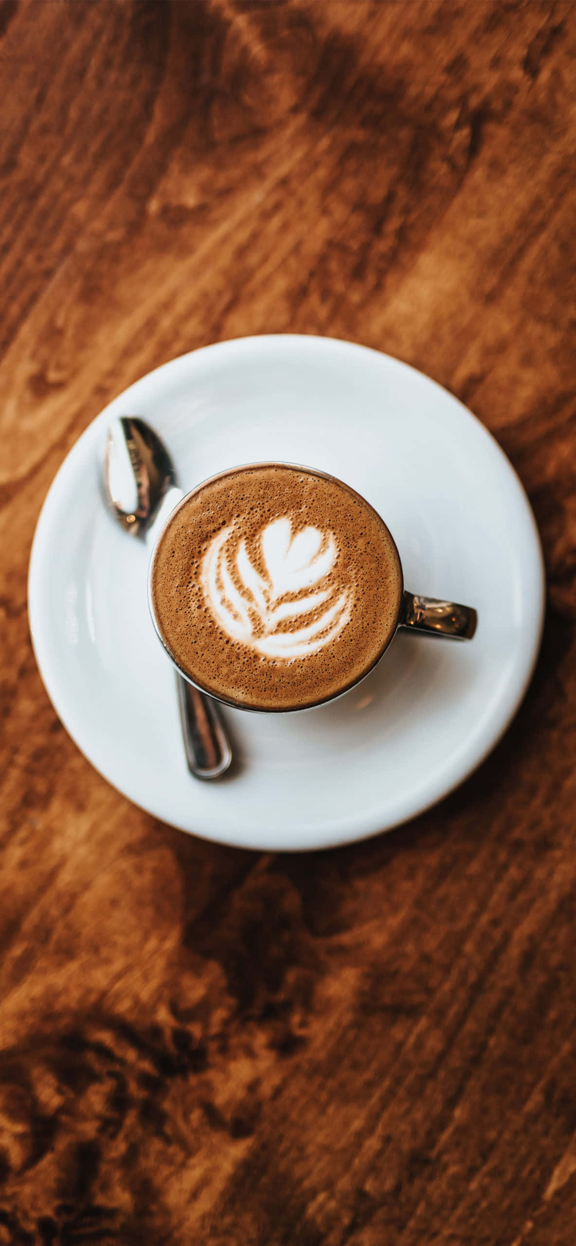 Coffee Aesthetic 桌布 壁紙 — 俯視圖：一個陶瓷杯中的心形葉奶泡藝術拿鐵，放在白色碟子上，旁邊有湯匙，置於深棕色木桌上。