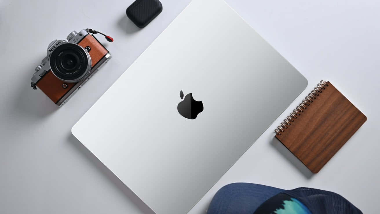 Mac 桌布 壁紙 — 俯視圖：一部銀色 MacBook、一部復古相機、木製筆記本和牛仔布帽整齊地排列在簡約的白色表面上。