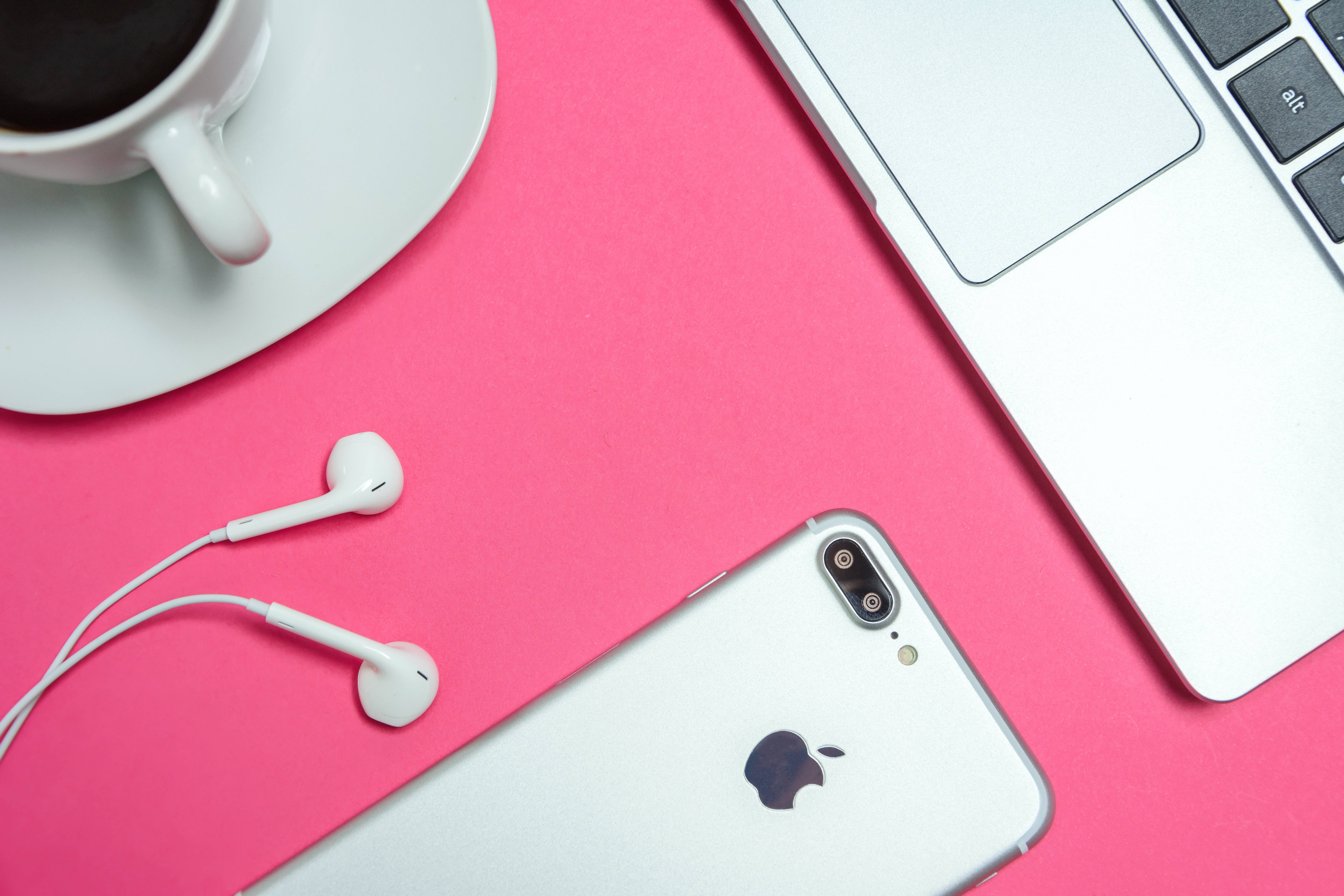 Hot Pink Aesthetic 桌布 壁紙 — 俯視圖：一部銀色iPhone、白色耳機和一杯咖啡，整齊擺放在鮮粉紅色表面上，旁邊有一部筆記型電腦。