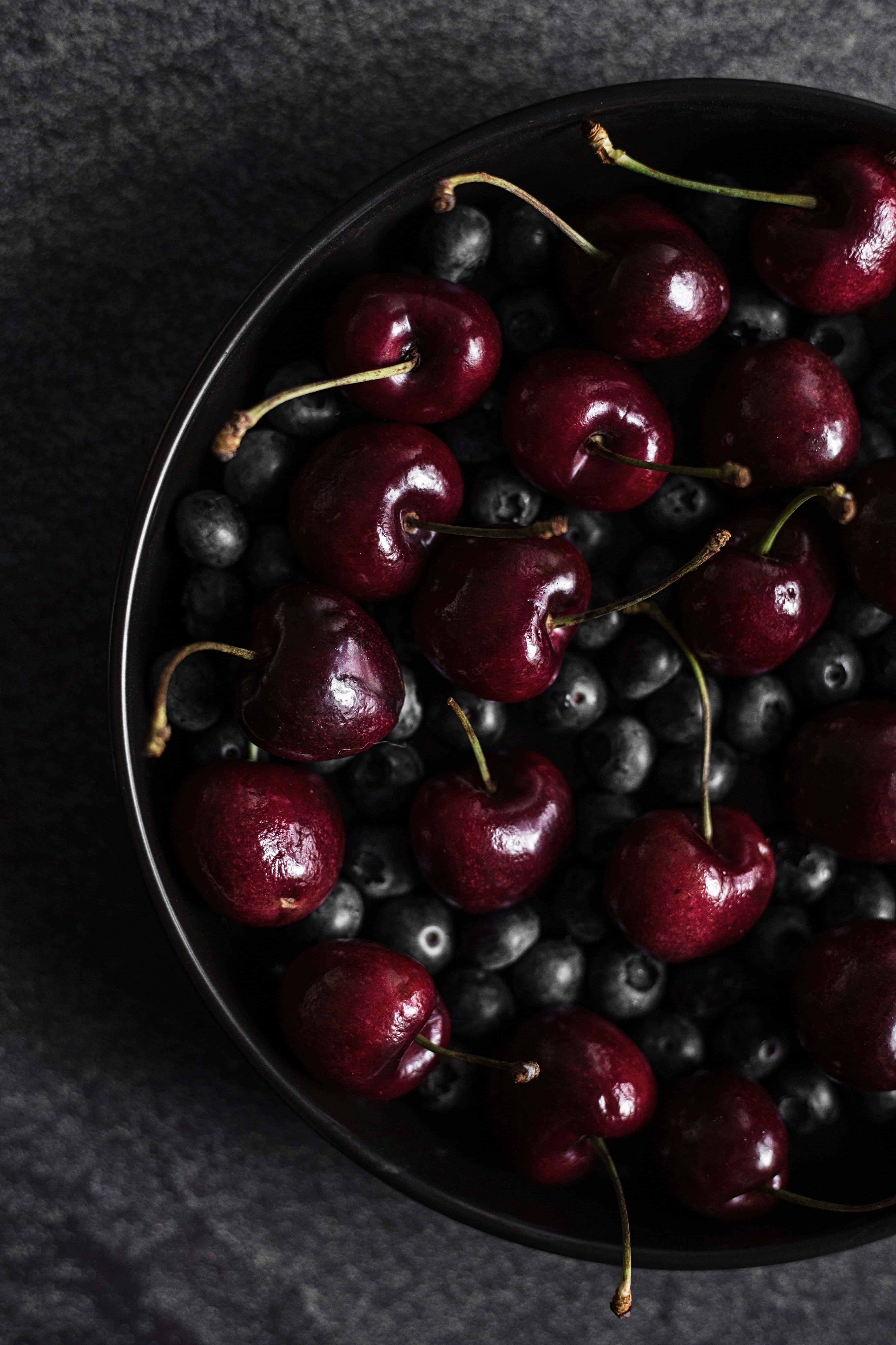 Black Cherry 桌布 壁紙 — 俯視圖，光澤紅櫻桃和藍莓依偎在啞光黑色碗中，背景為深色紋理表面。