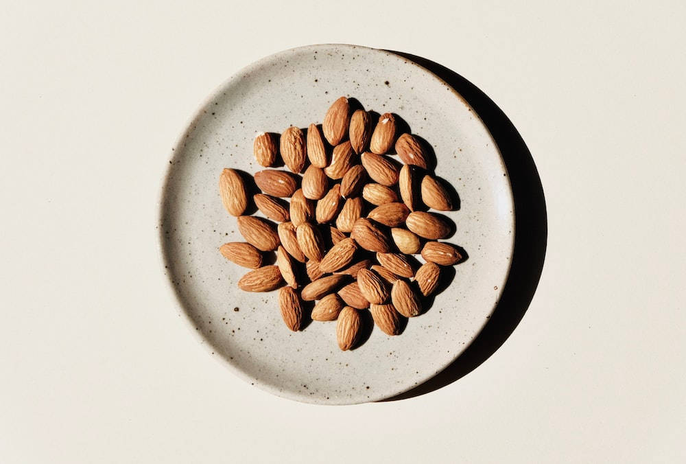 Almond 桌布 壁紙 — 俯視圖，杏仁散落在有斑點的陶瓷盤上，在純色背景上投下銳利的陰影。