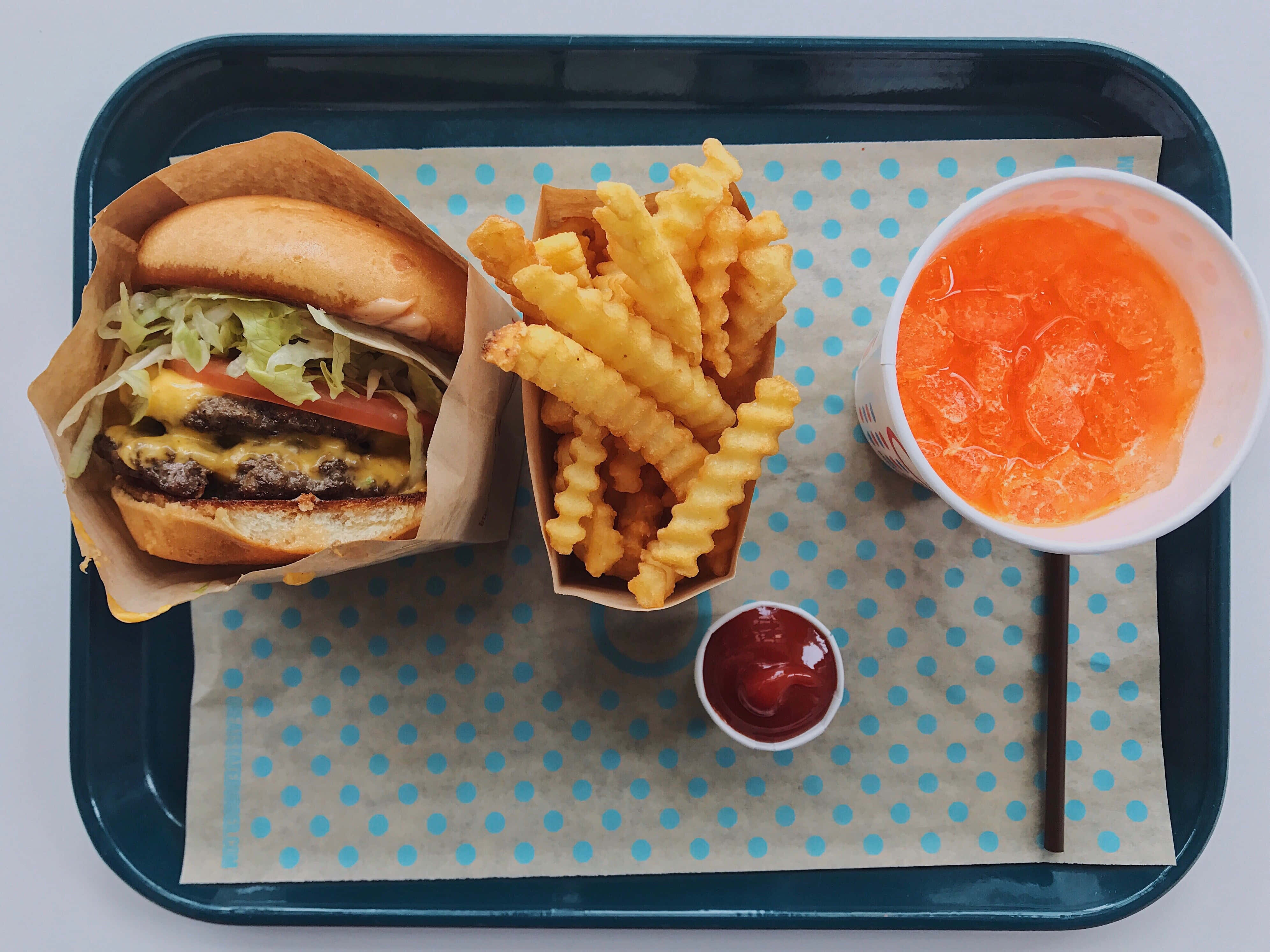 fast food 桌布 壁紙 — 俯視圖：雙層起司漢堡、波浪薯條和柳橙飲料放在藍色托盤上，托盤鋪有圓點紙。