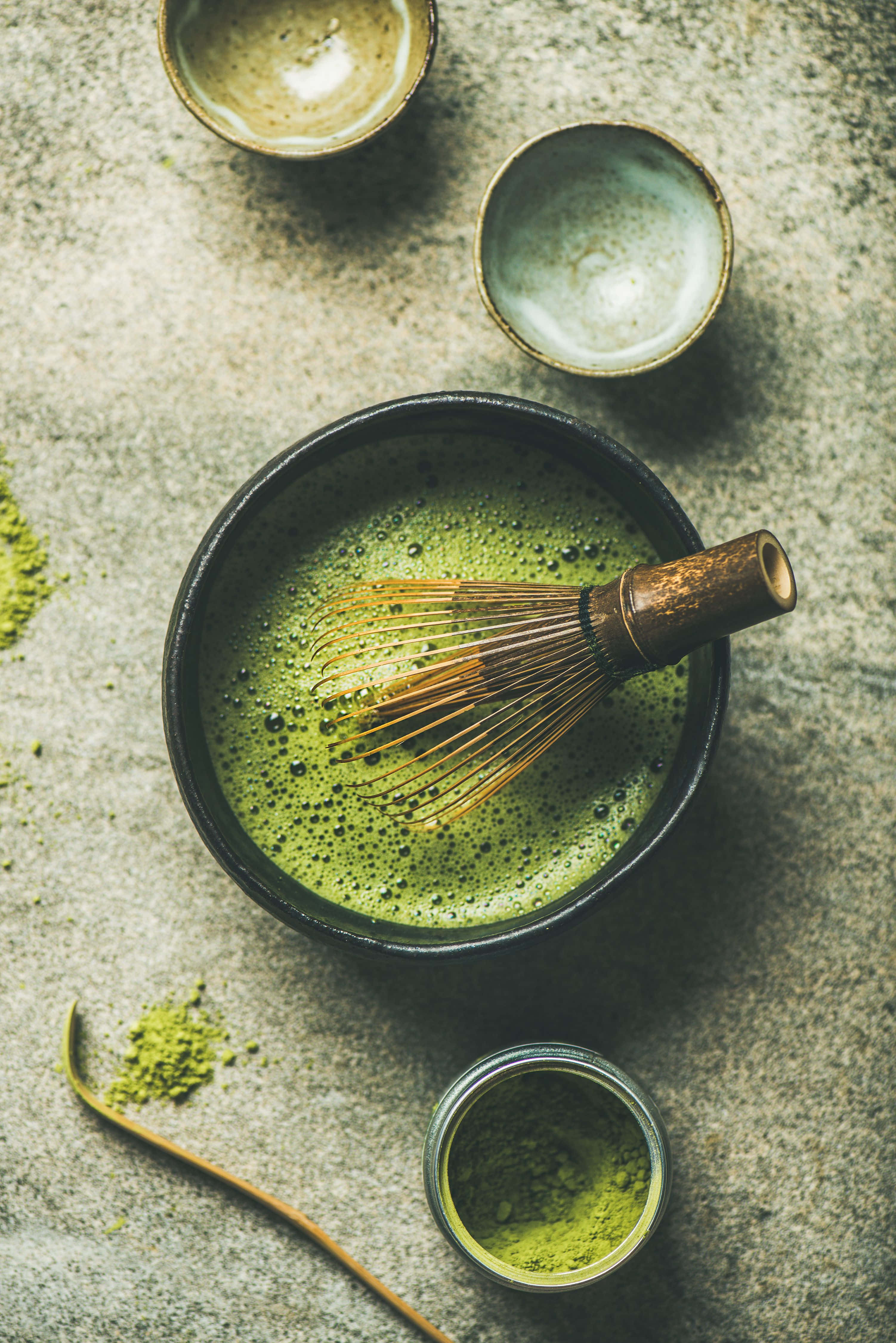 Matcha 桌布 壁紙 — 俯視圖，黑色碗中盛有鮮綠色抹茶，竹筅置於其中，周圍有陶瓷杯和粉末散落在有紋理的石面上。