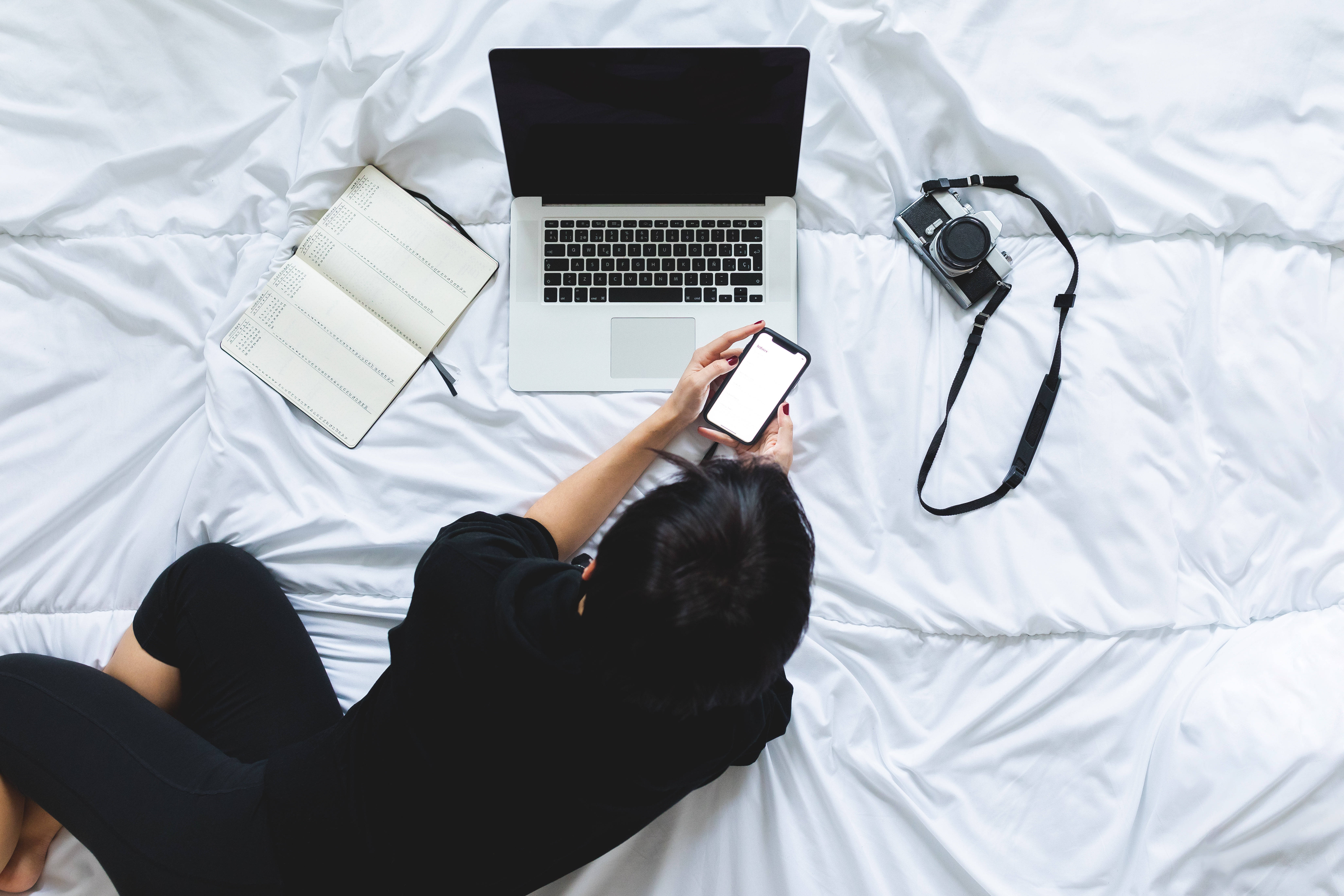 Laptop Desktop 桌布 壁紙 — 俯視角度，一名穿黑色衣服的人躺在白色床上使用手機，附近有筆記型電腦、筆記本和相機。
